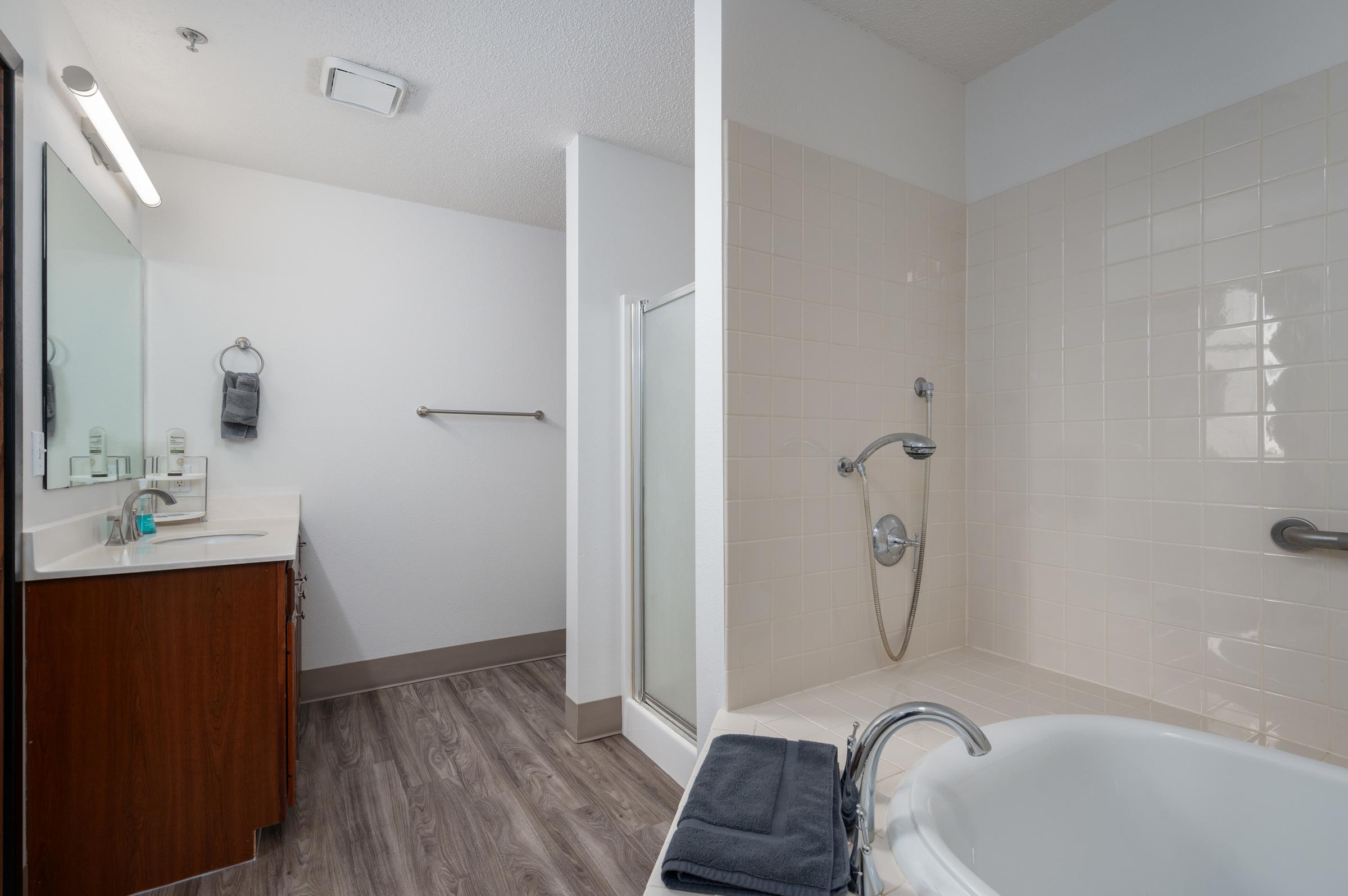 A modern bathroom featuring a double sink vanity with a large mirror, a walk-in shower with glass door, and a separate bathtub. The walls are tiled in light colors, and there is a towel rack on the wall. Wood-look flooring adds warmth to the space.