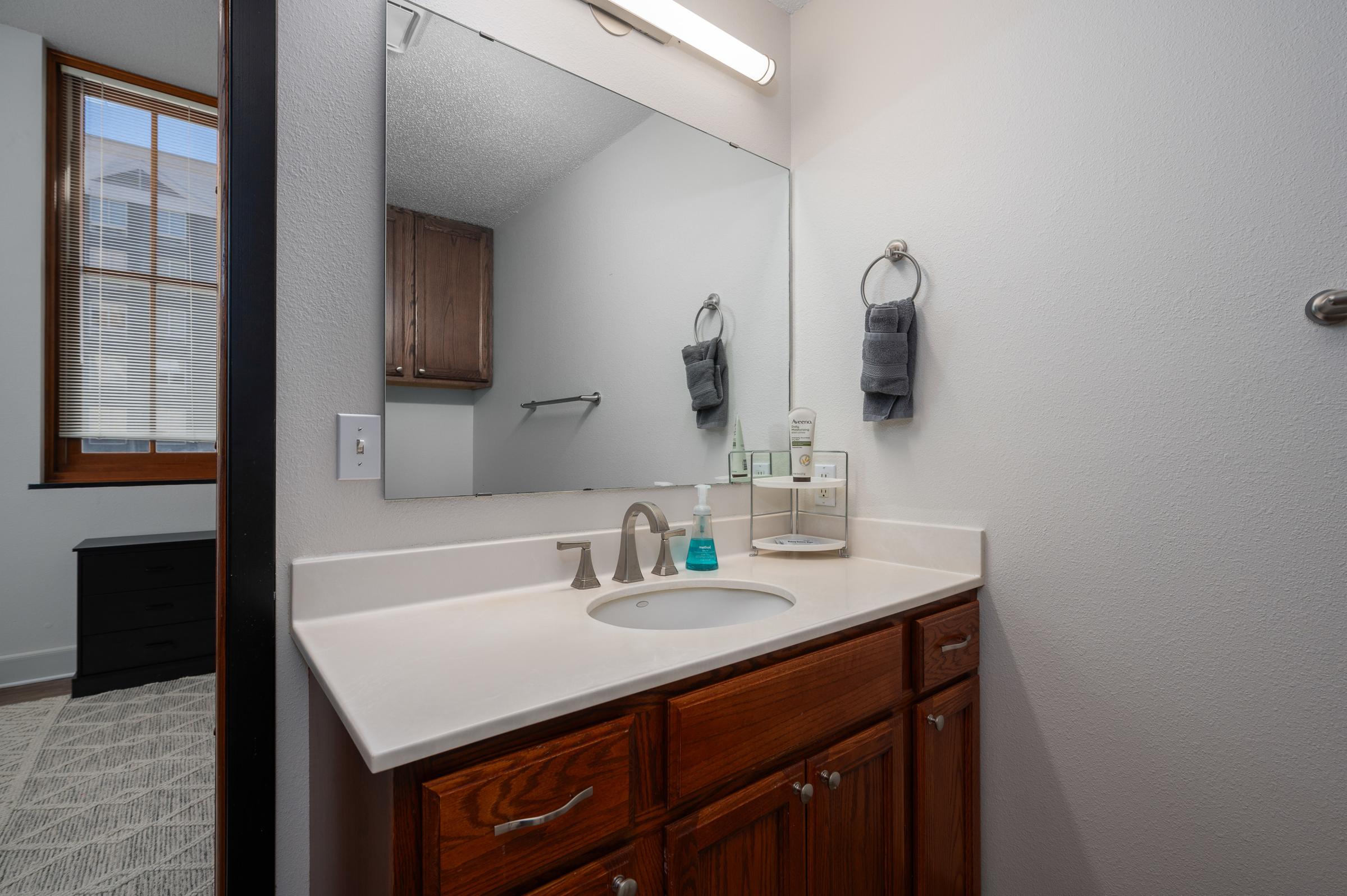 A modern bathroom featuring a wooden vanity with a sink and faucet, a large mirror above, and neatly hung gray towels. The space is well-lit, showcasing a clean design with neutral colors. In the background, a dark wooden drawer unit is visible, and natural light filters through a window.