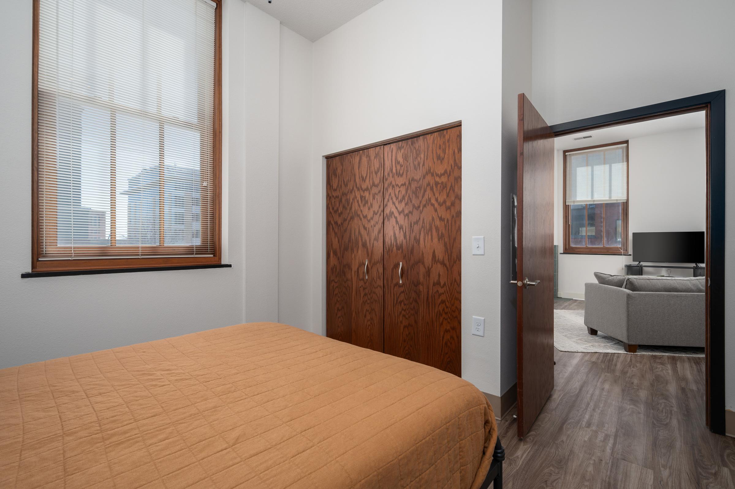 A cozy bedroom featuring a warm-toned bedspread, a wooden wardrobe, and a doorway leading to a living area. Natural light streams through large windows, highlighting the neutral decor and modern furnishings, including a grey sofa and a TV in the adjacent space.
