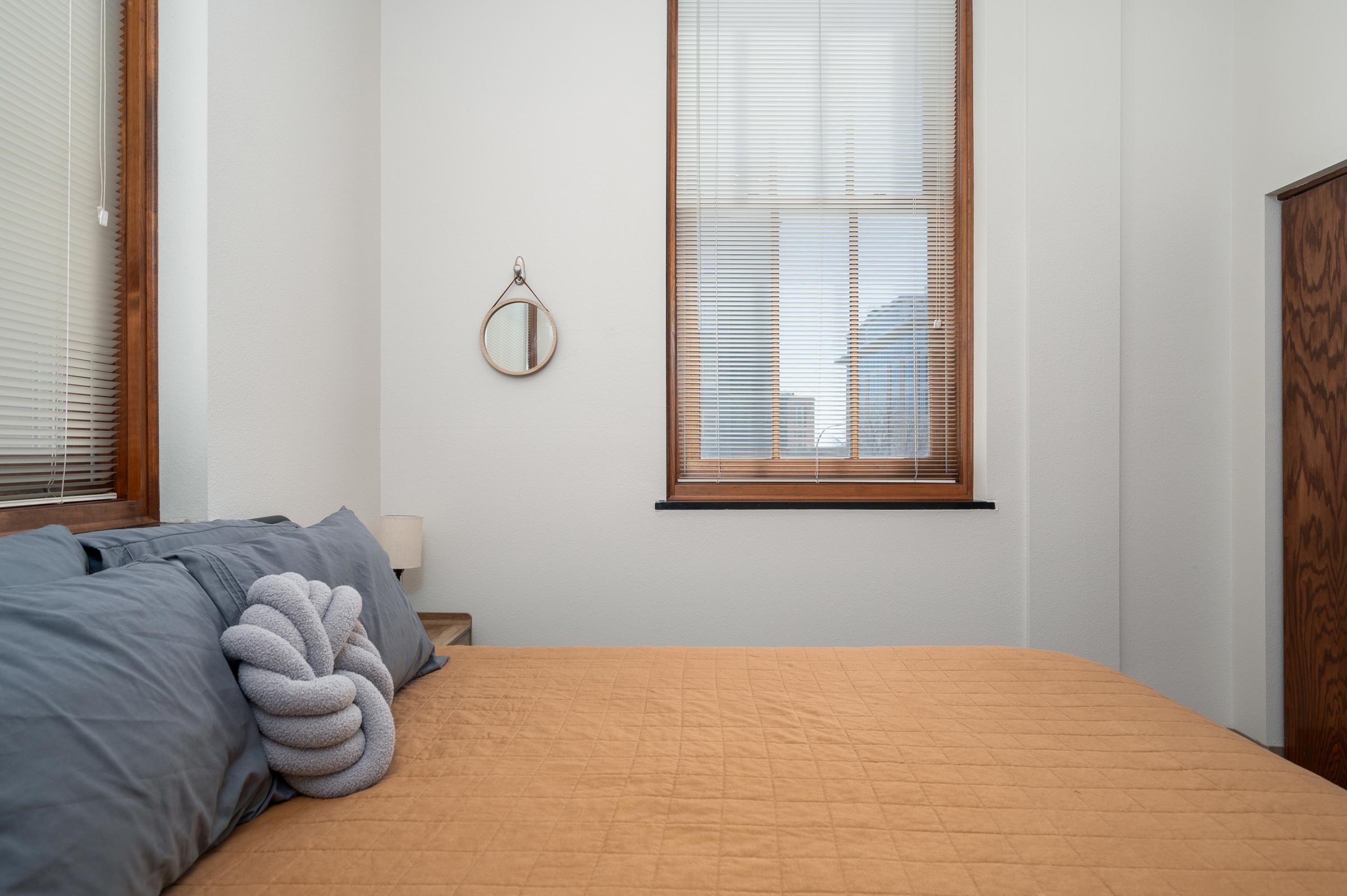 A cozy bedroom featuring a neatly made bed with an orange quilt and gray pillows. A decorative gray knot-shaped pillow rests on the bed. A small round mirror hangs on the wall, and there's a window allowing natural light to filter in, casting soft shadows on the walls.