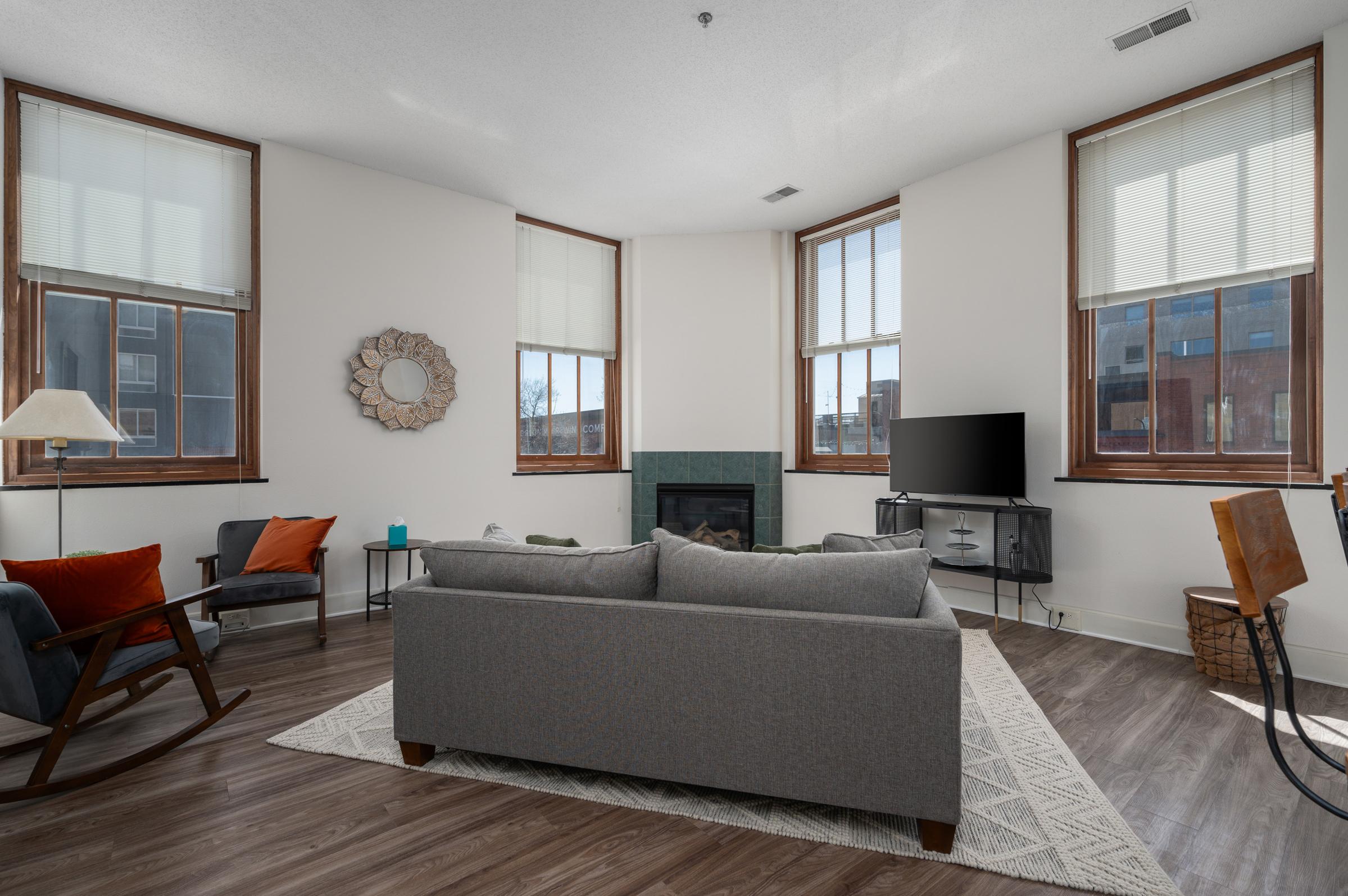 A cozy living room featuring a gray sofa, two chairs with orange cushions, and a decorative mirror on the wall. Natural light fills the space from large windows with white blinds. A fireplace is visible in the corner, and a television is mounted on a stand, adding to the inviting atmosphere.