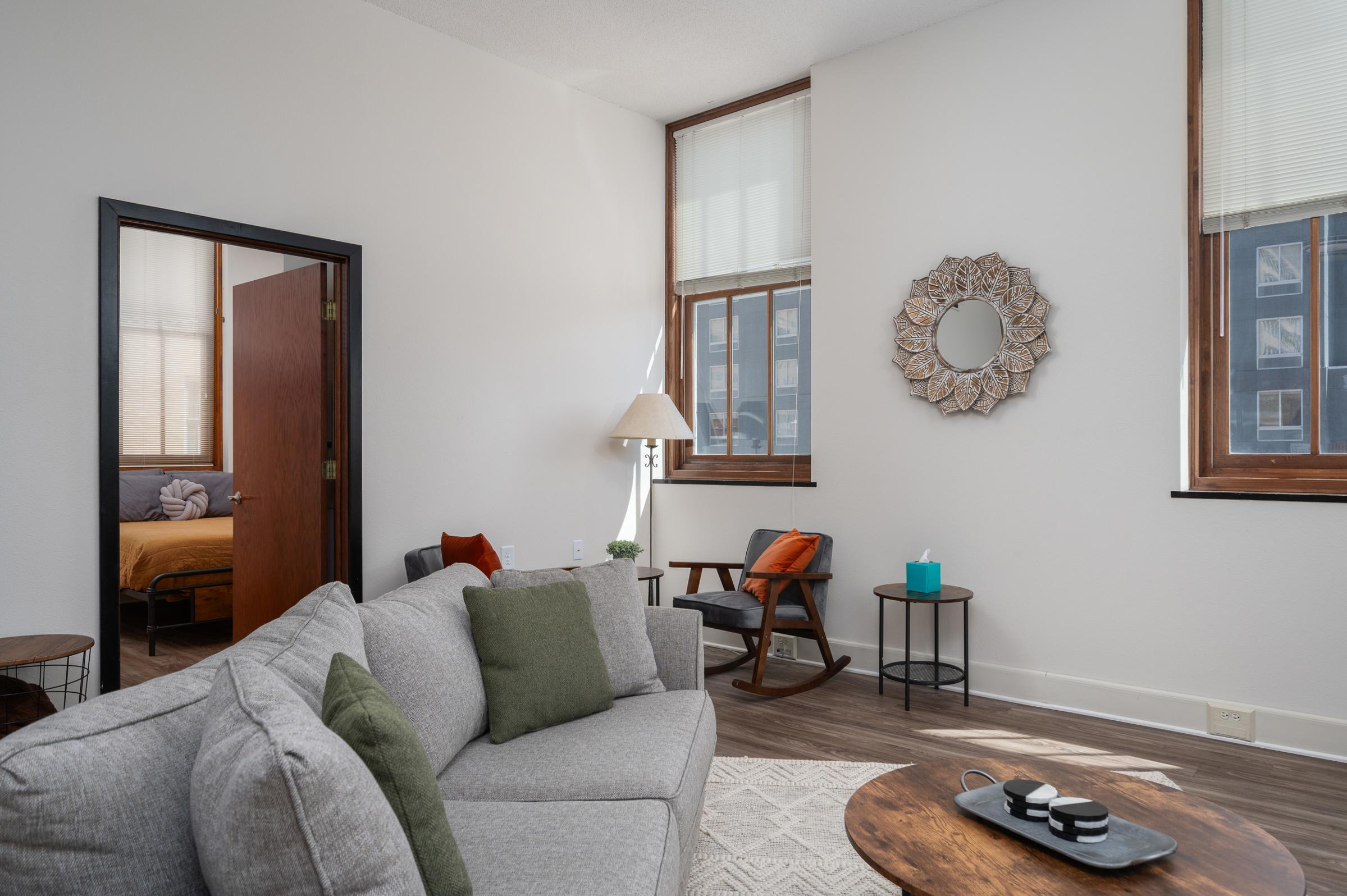 A cozy living room featuring a light gray sectional sofa with green and orange cushions, a round wooden coffee table, and a rocking chair. Light floods in through two large windows with blinds. A decorative mirror hangs on the wall, and a doorway leads to a bedroom in the background.