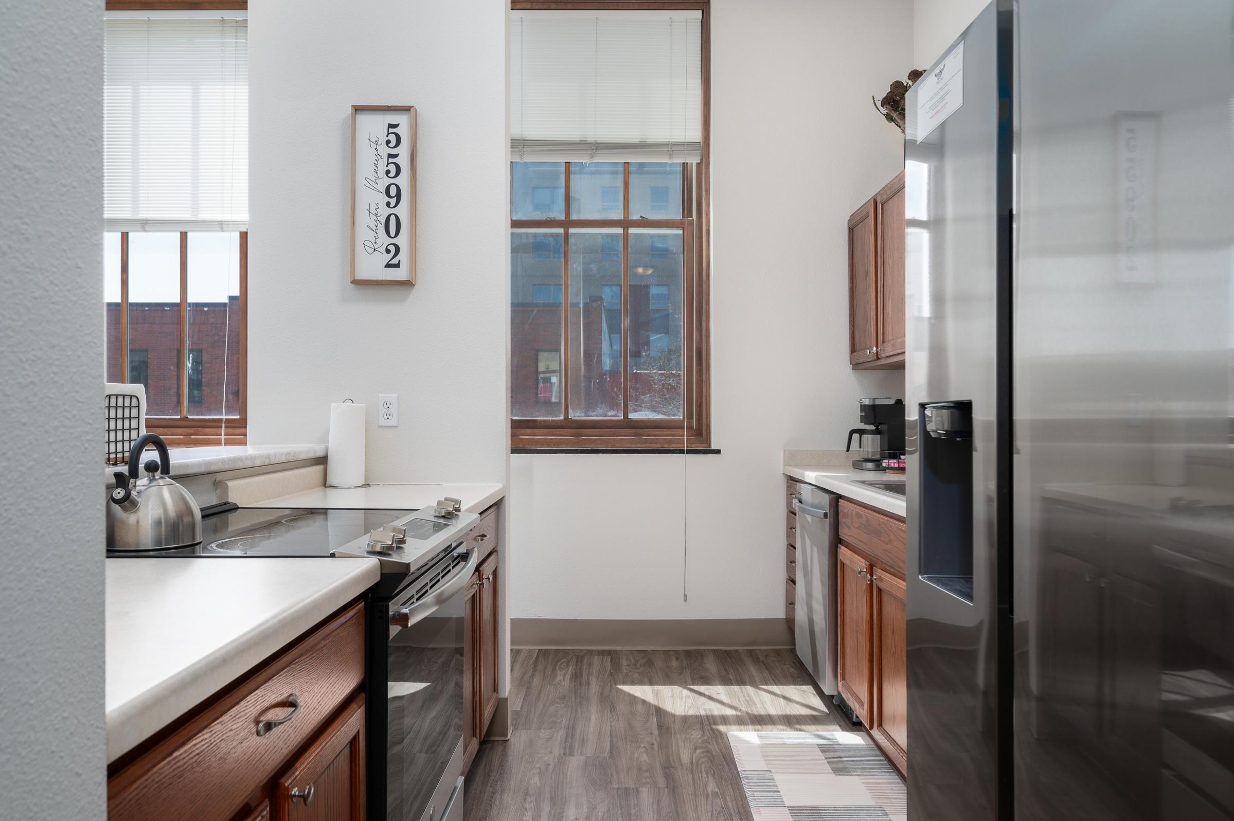 A well-lit kitchen featuring wooden cabinets, stainless steel appliances, and a countertop with a kettle and paper towel. Large windows allow natural light, and a small rug is placed on the floor. A wall sign displays a number, adding a decorative touch to the space.