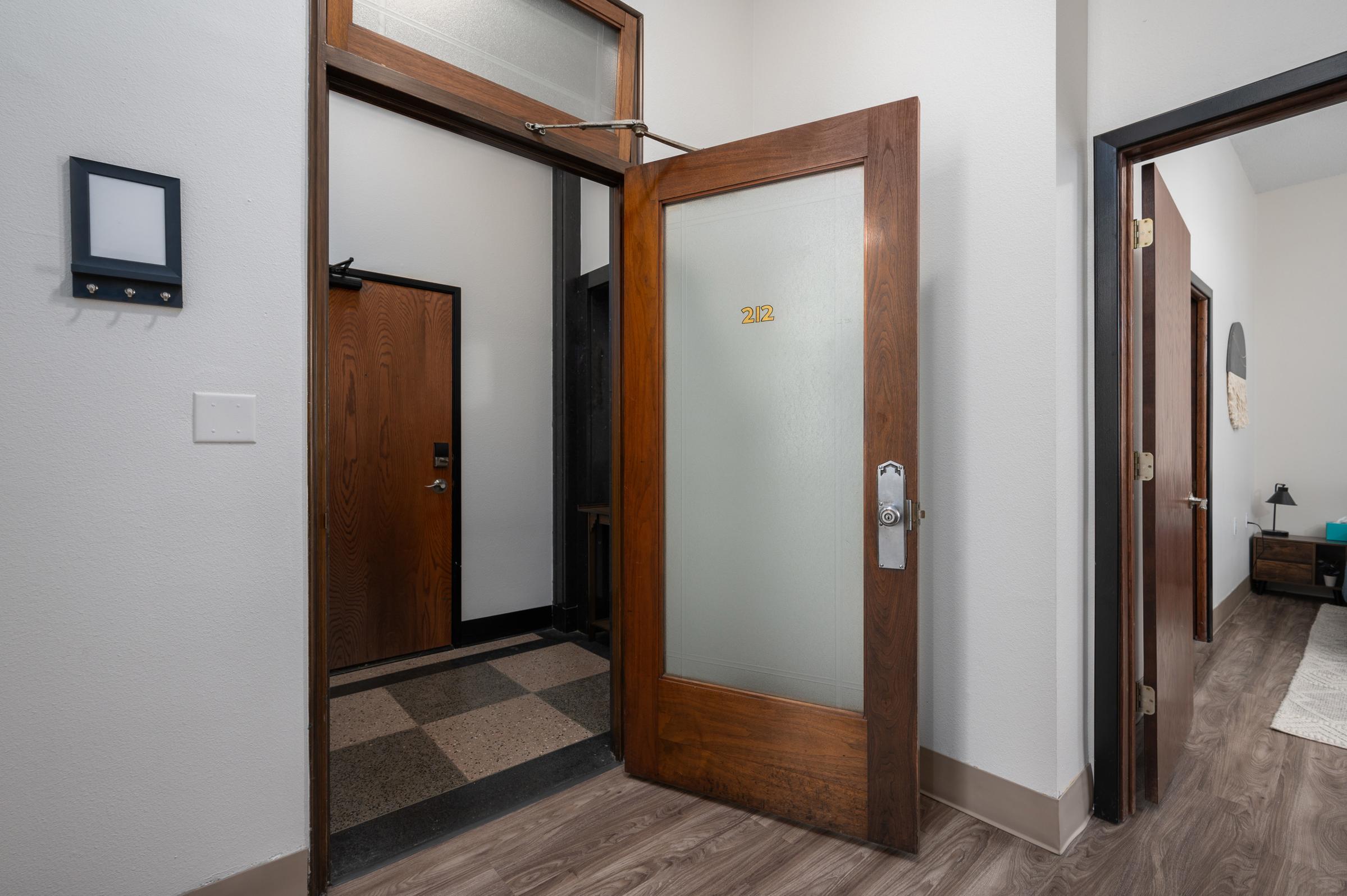 Interior view of a hallway featuring an open wooden door with a frosted glass panel. Another door is partially visible on the left. The floor has a checkered pattern, and the walls are painted white. A digital keypad is mounted on the wall near the entrance. Modern lighting and decor elements can be seen.