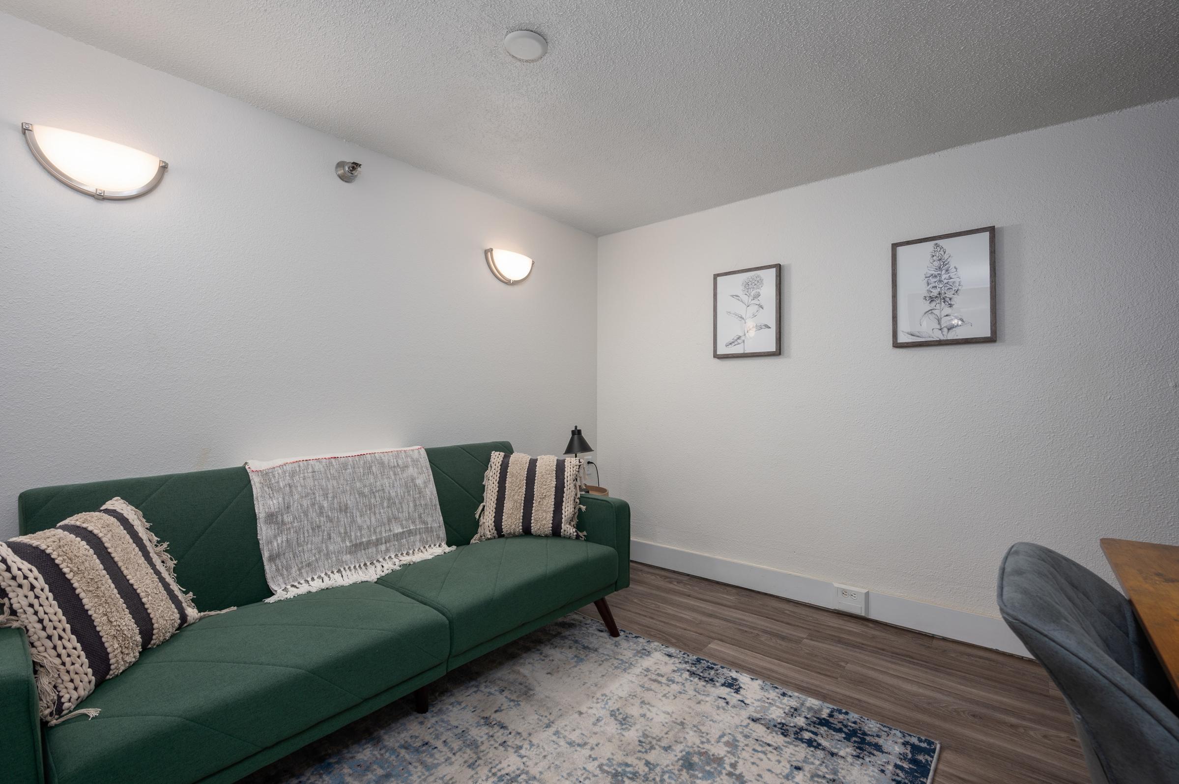 A cozy living room featuring a green sofa with decorative pillows, two framed botanical prints on the white wall, and a small lamp on a side table. The flooring is wood-like, and a light blue and white area rug adds a touch of color to the space.
