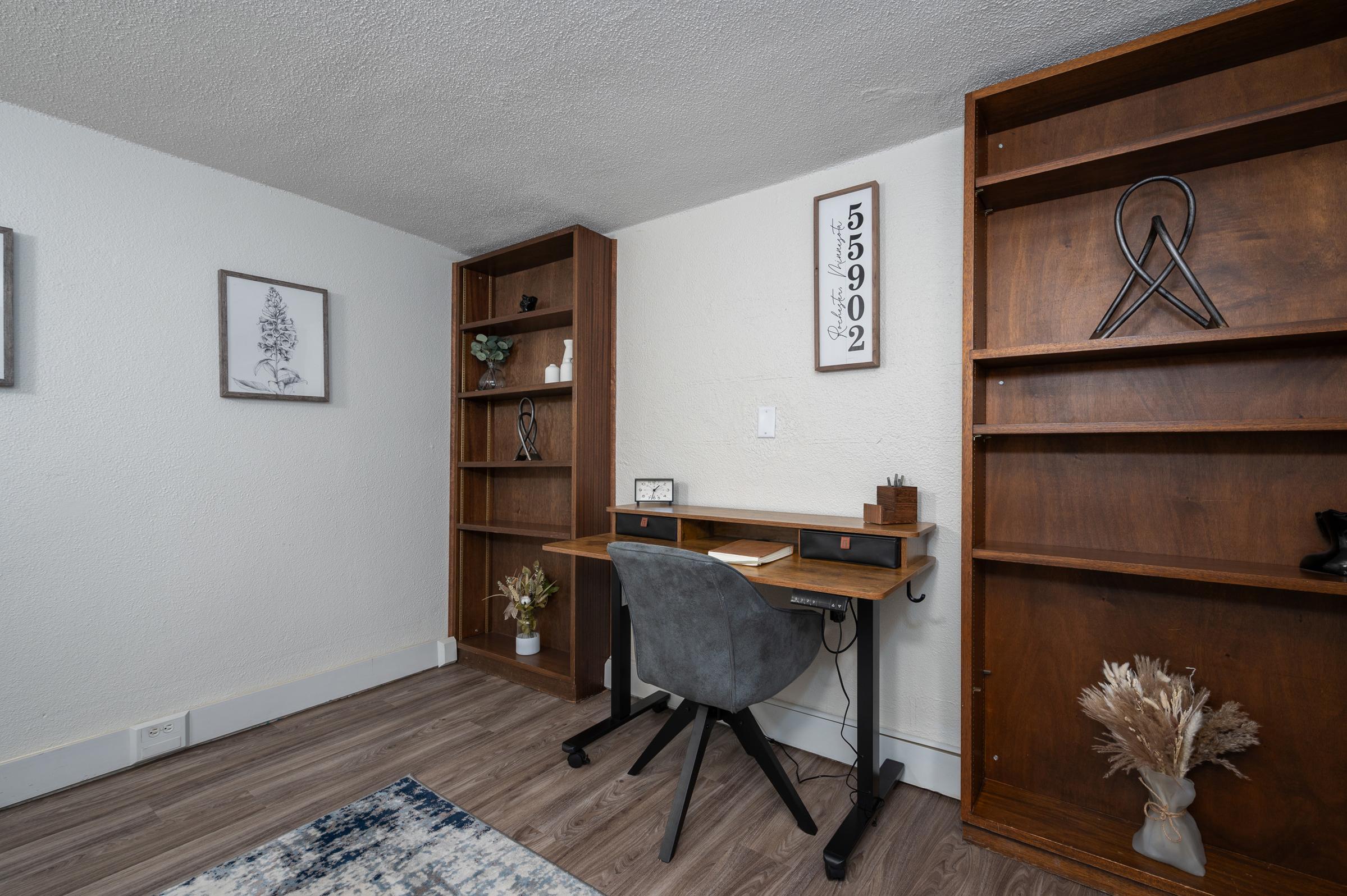 A cozy home office setup featuring a wooden desk with a modern chair, surrounded by two wooden bookshelves. One shelf holds decorative items and a small plant, while the wall displays framed art. The flooring is light wood, and the walls are painted white, creating a bright and inviting workspace.