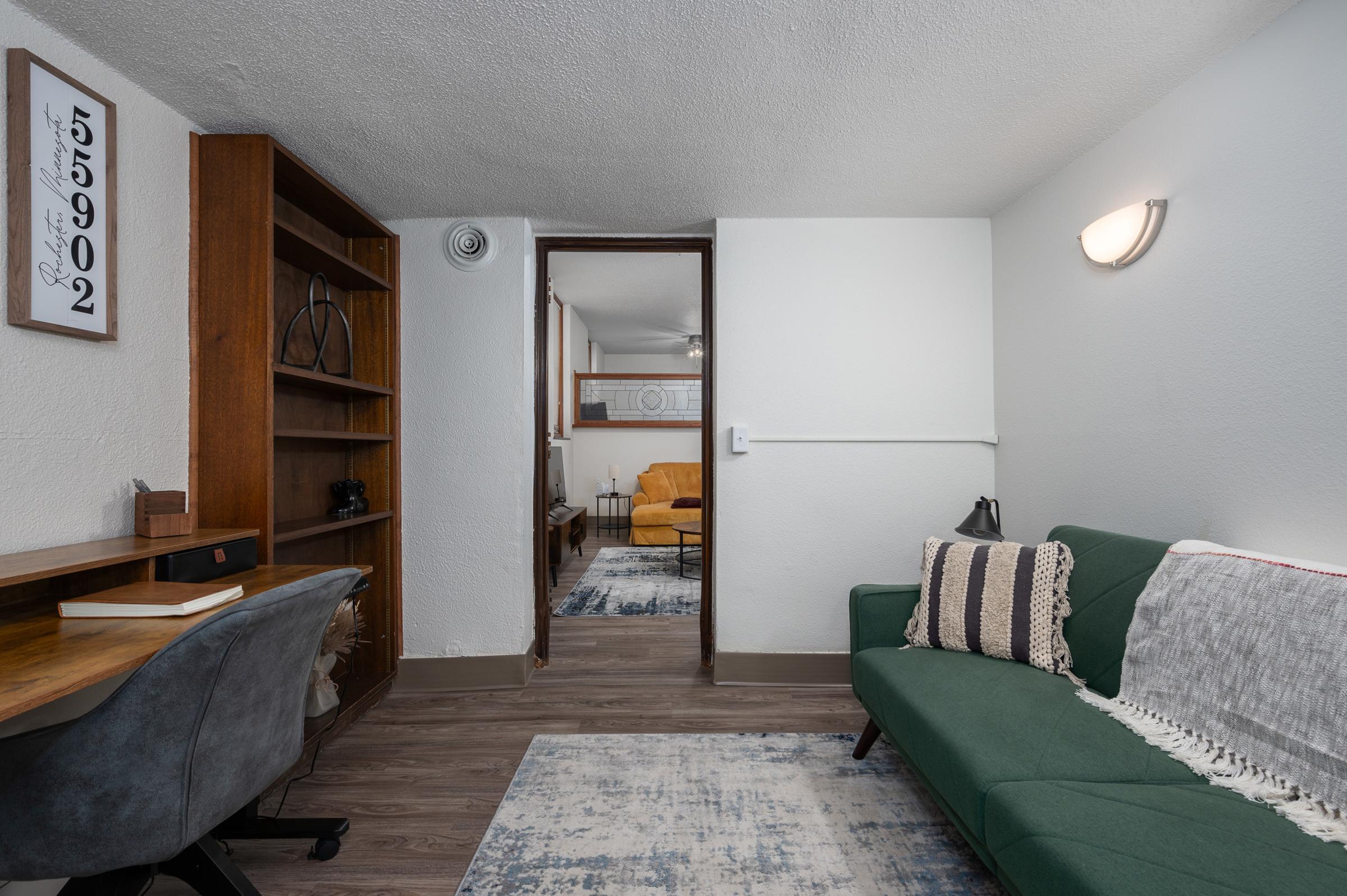 A cozy living space featuring a green sofa with decorative pillows, a wooden desk, and a bookshelf. Light-colored walls and a rug enhance the inviting atmosphere. A doorway in the background leads to another room with a yellow chair, adding a pop of color. Soft lighting creates a warm ambiance.