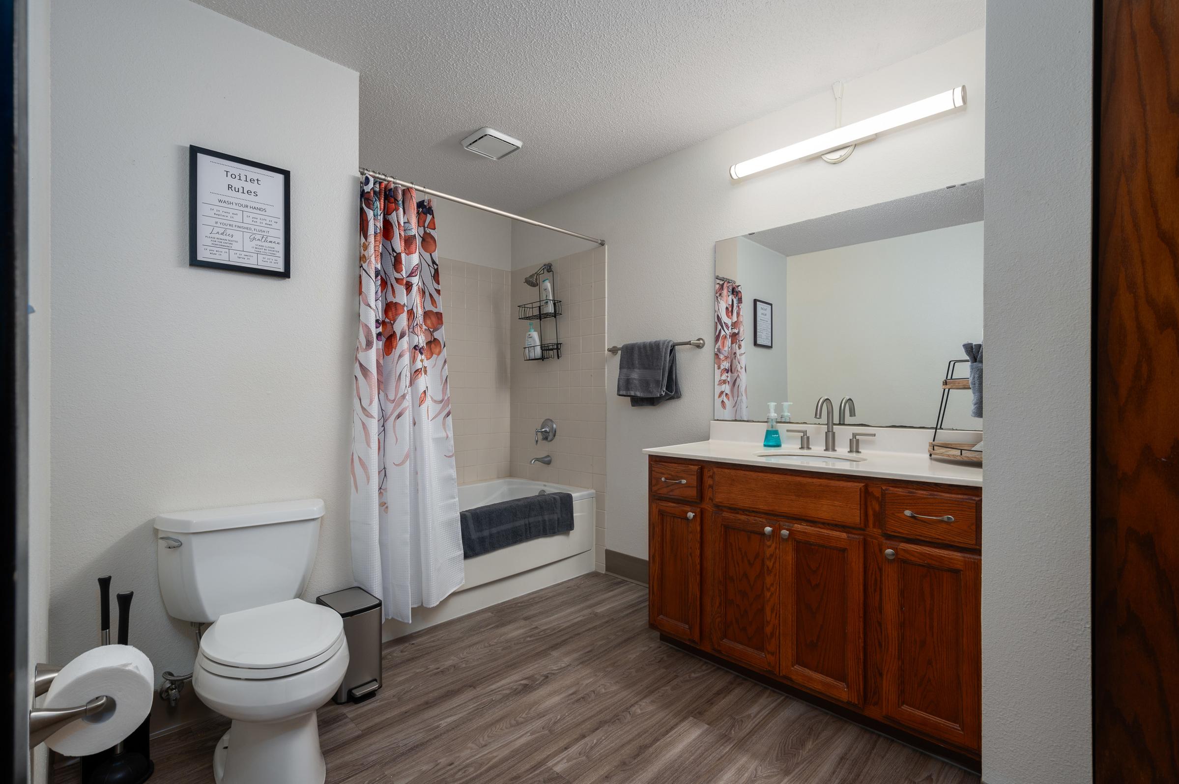 A modern bathroom featuring a bathtub with a shower curtain adorned with floral designs, a wooden vanity with a sink, a mirror above the vanity, a toilet, and a small trash bin. The floor is wood-like, and the walls are painted a light color. A towel is hanging on a rack, and there is overhead lighting.