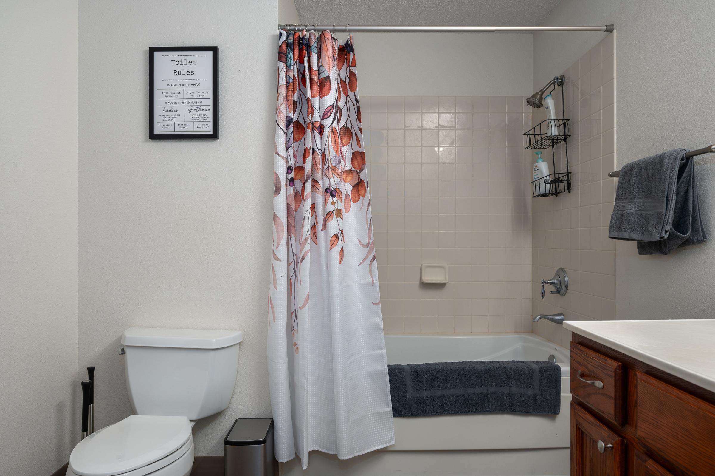 A clean bathroom featuring a white toilet, a bathtub with a floral shower curtain, and a towel rack. The walls are light-colored, and there's a framed sign with "Toilet Rules" on the wall. A gray towel is neatly placed on the rack, and a small trash can is in the corner.