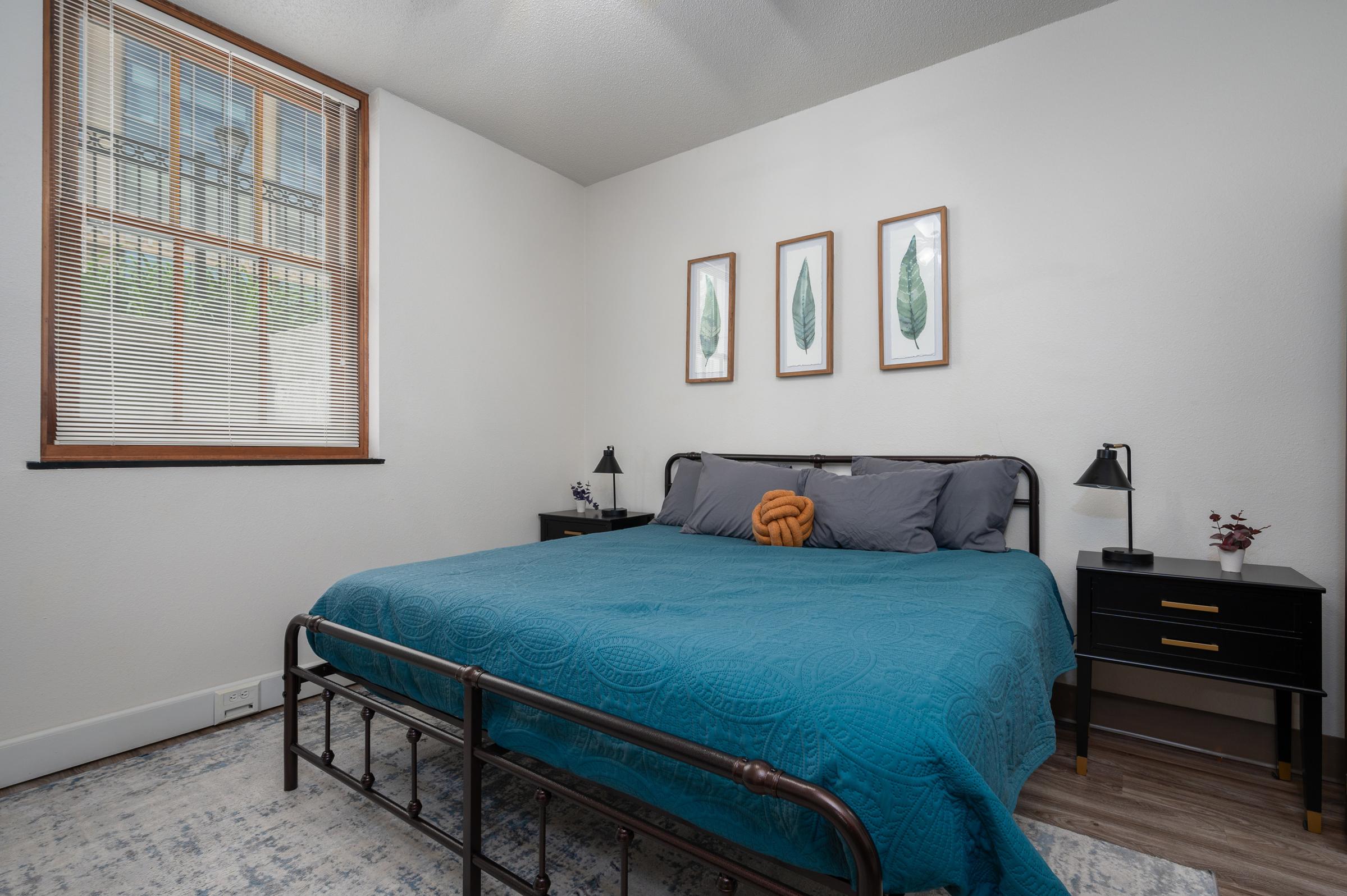 A cozy bedroom featuring a metal bed frame with a teal quilt, accent pillows, and nightstands with lamps. Three framed leaf prints hang on the wall above the bed, and a window provides natural light. The room has light wood flooring and a neutral color scheme with minimal decor.