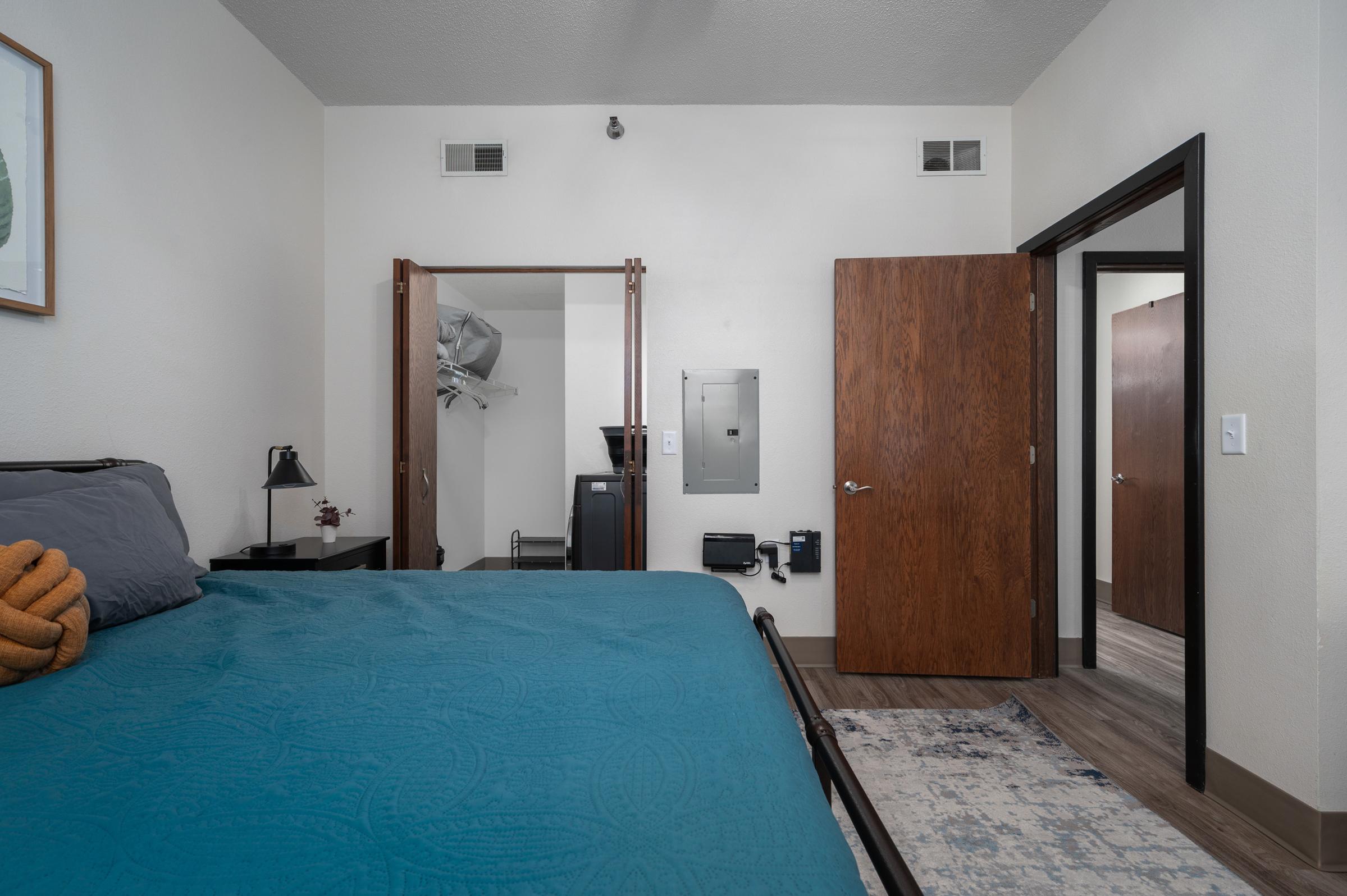 A cozy bedroom featuring a blue bedspread, a lamp on a nightstand, and wooden doors leading to an adjacent area. One door reveals a closet, while the other leads to a bathroom or hallway. The room has neutral wall colors and includes a decorative rug on the floor.