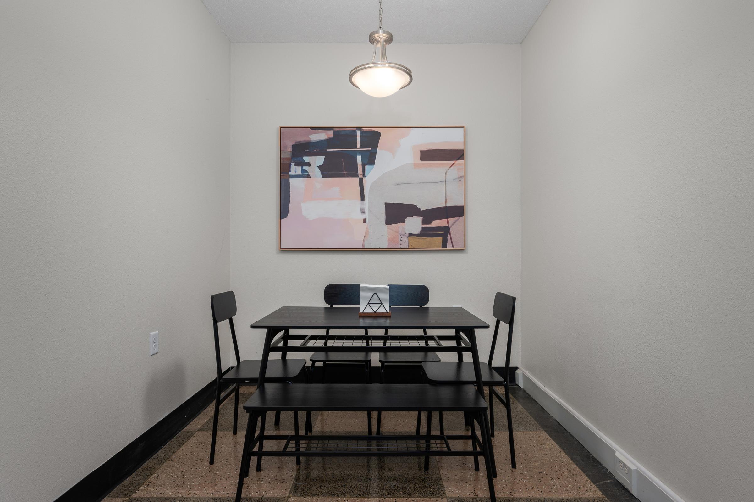 A modern, minimalist dining area featuring a black table and chairs. A piece of abstract wall art with soft pinks and blues hangs above the table, which is adorned with a small decorative item in the center. The space has a neutral wall and a pendant light overhead, giving it a clean, contemporary look.