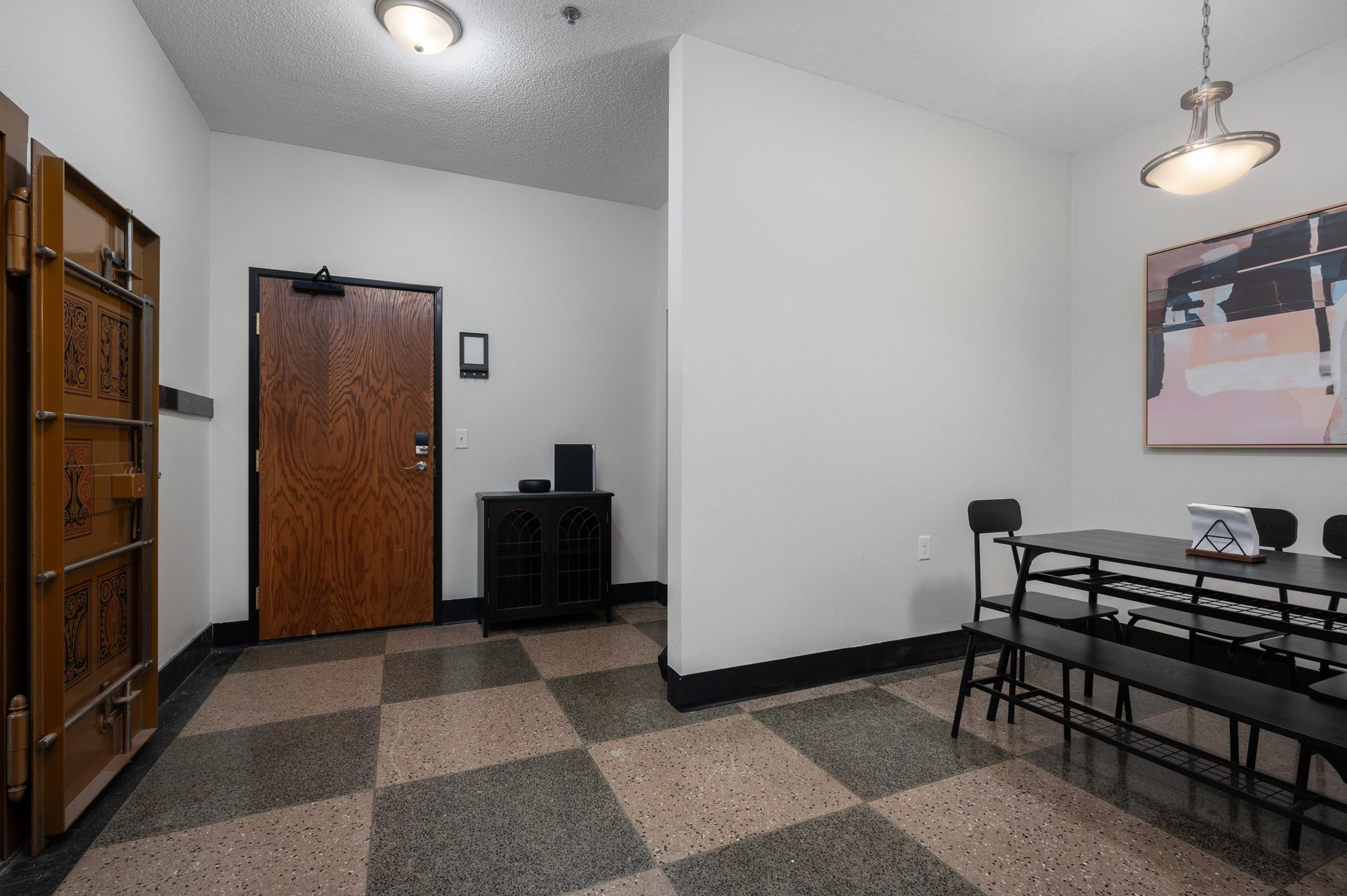 A well-lit interior space featuring a wooden door with a visible keycard entry, a small black cabinet, and a dining table with black chairs. The floor has a checkerboard pattern with light and dark tiles, and a decorative wall art piece hangs nearby. Soft overhead lighting creates a warm atmosphere.