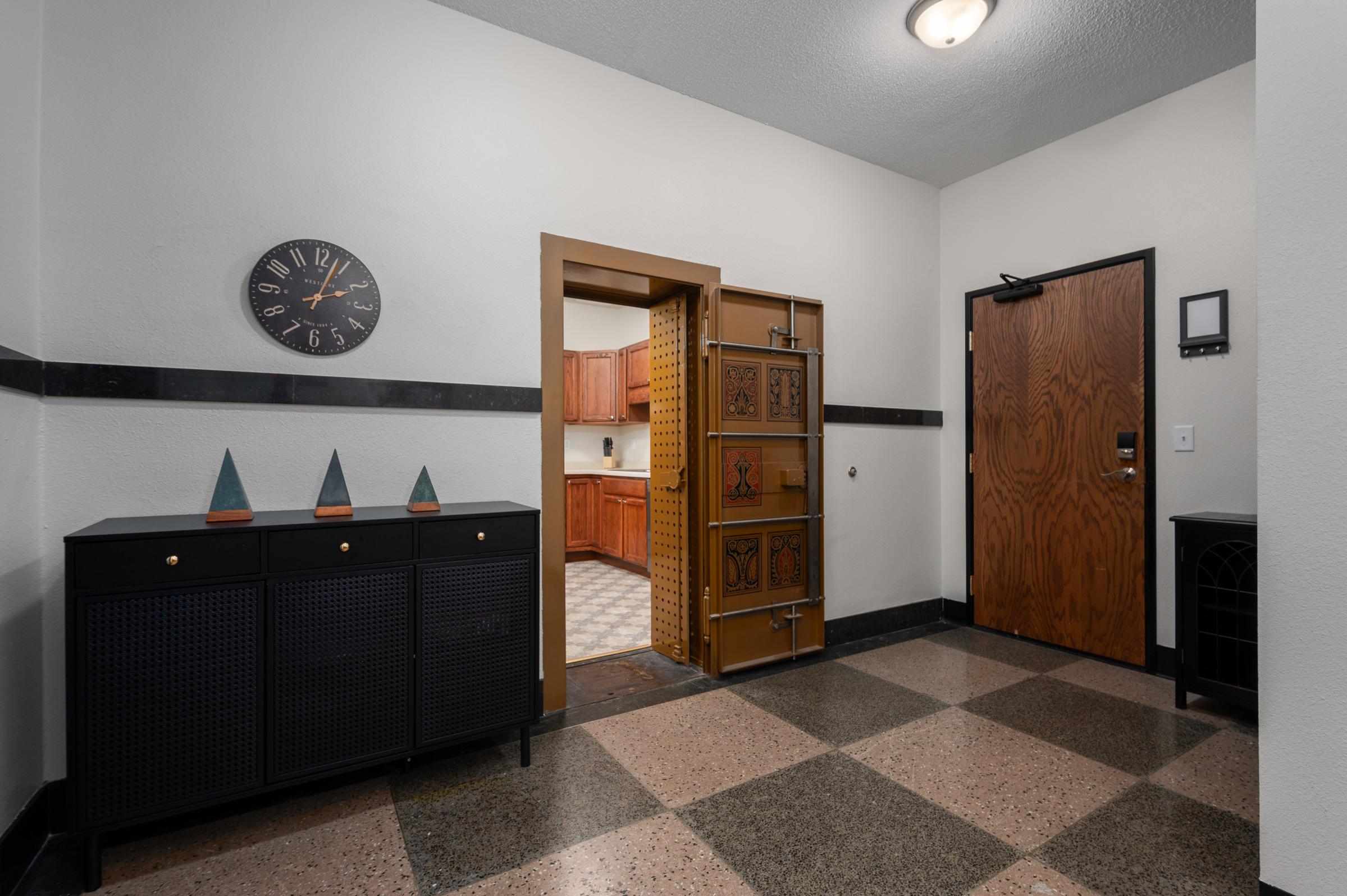 Interior view of a hallway featuring a decorative black and gold entry door, a wooden door leading to a kitchen, a wall clock, and a sideboard with three decorative objects. The floor has a checkered pattern, and the walls are painted in neutral tones.