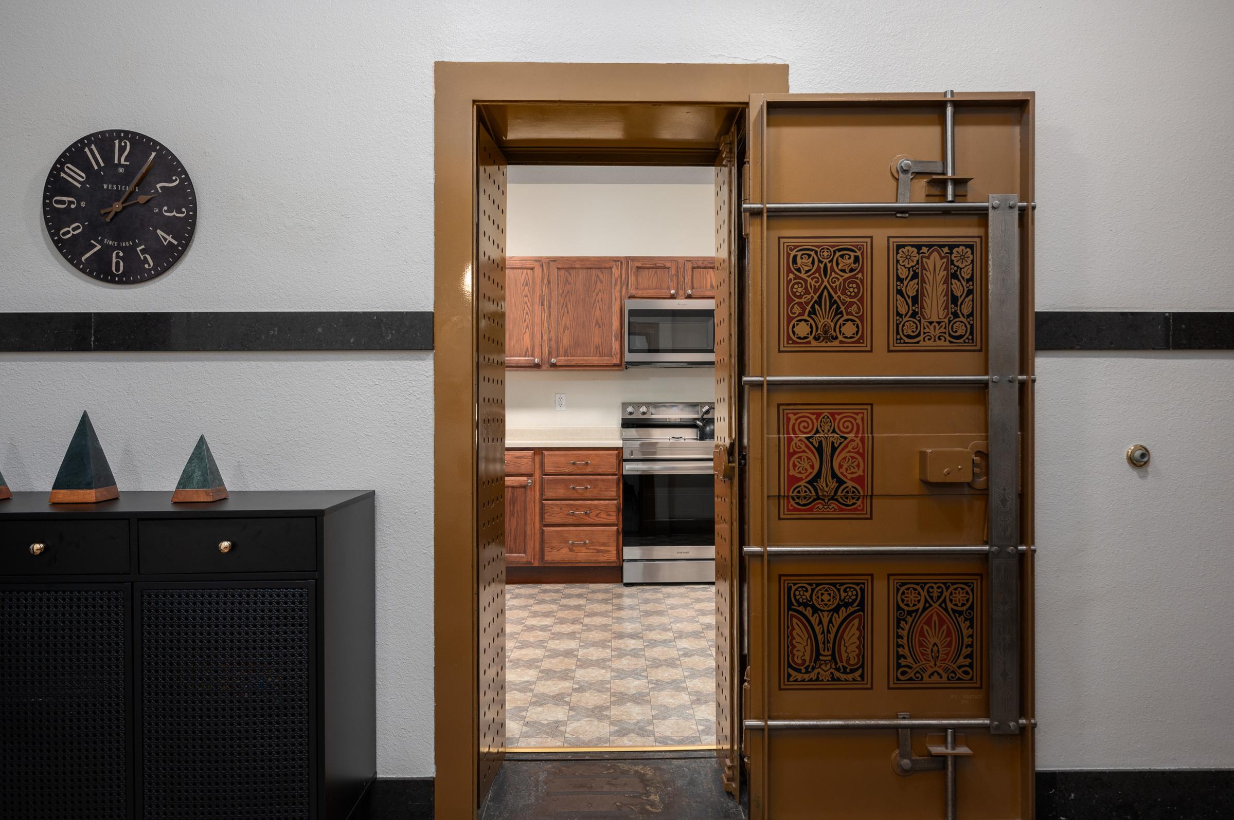 A view through a secure door leading to a kitchen. The kitchen features wooden cabinets, modern appliances, and patterned tile flooring. A clock is mounted on the wall beside the door, and decorative triangular items are placed on a cabinet in the foreground, adding a touch of decor to the entryway.