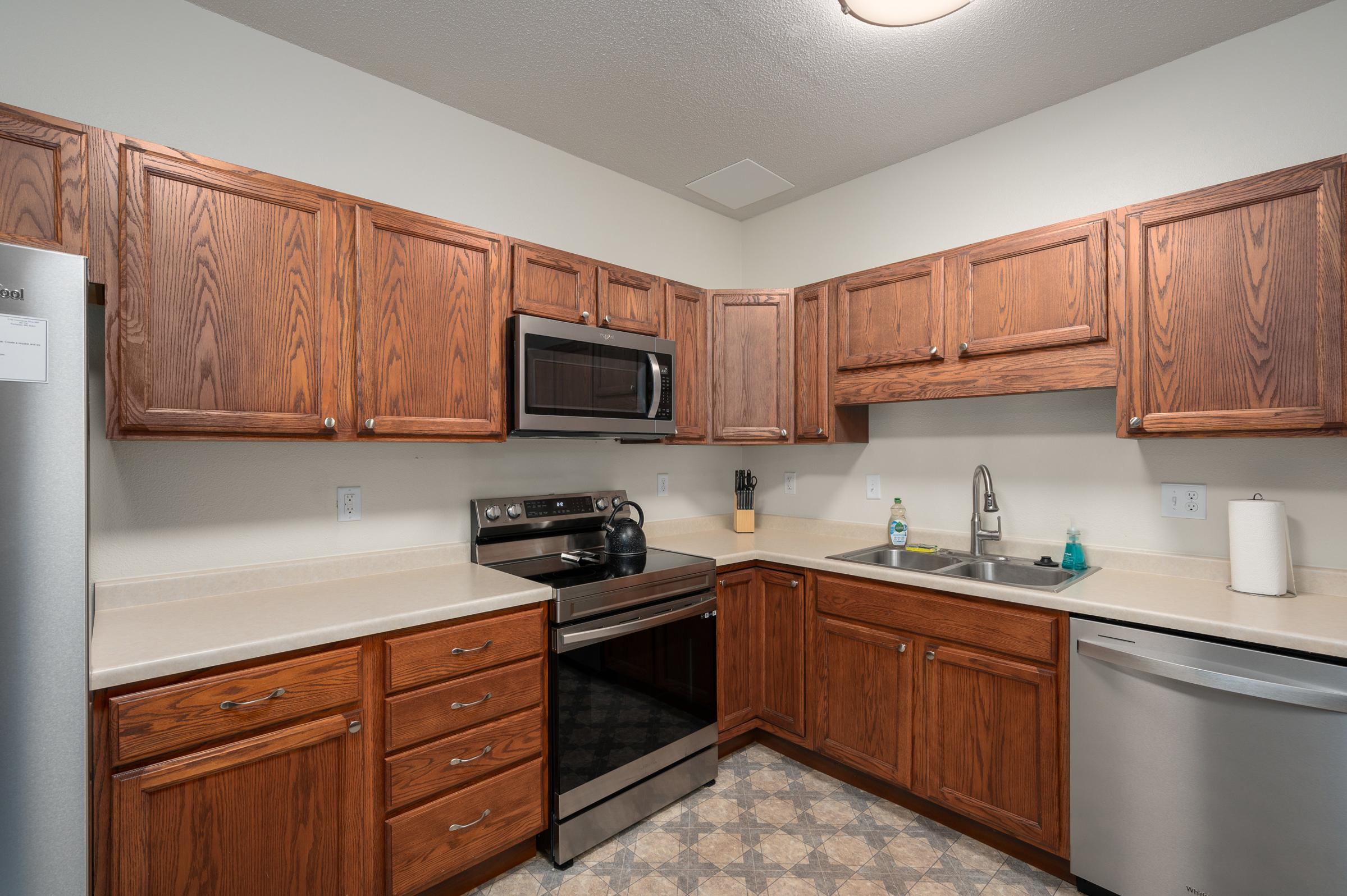 A well-lit modern kitchen featuring wooden cabinetry, an electric stove, microwave, and stainless steel appliances. The countertop is beige with a sink and dish soap visible. The floor has a tiled pattern, and there are kitchen utensils and a soap dispenser visible on the countertop.