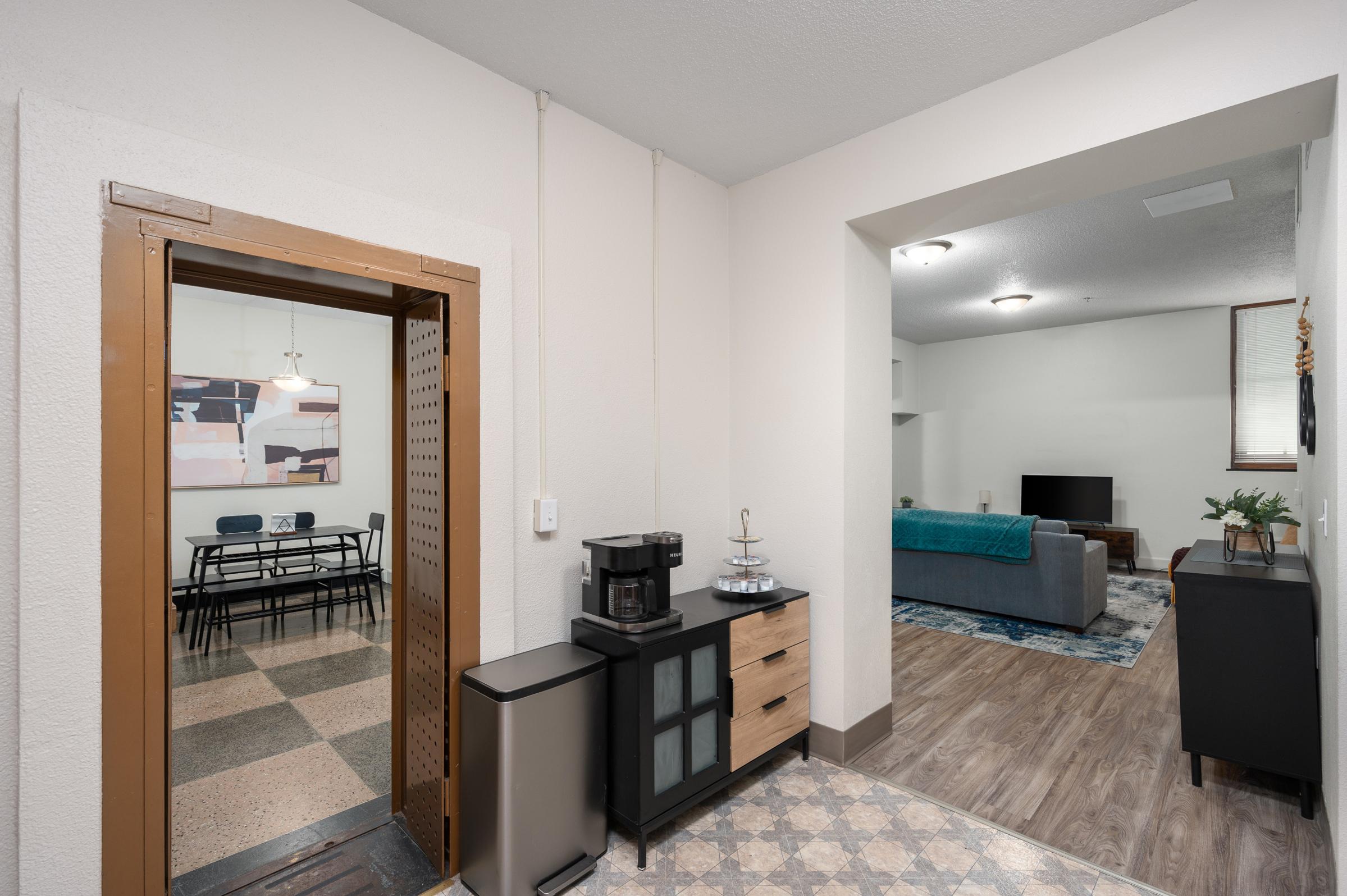 View of an apartment entrance transitioning into a living area. On the left, there's a kitchen area with a coffee maker and a small fridge. The right side shows a living room with a couch, TV, and a dining table visible in the background. The walls are painted light colors, creating a bright and welcoming atmosphere.