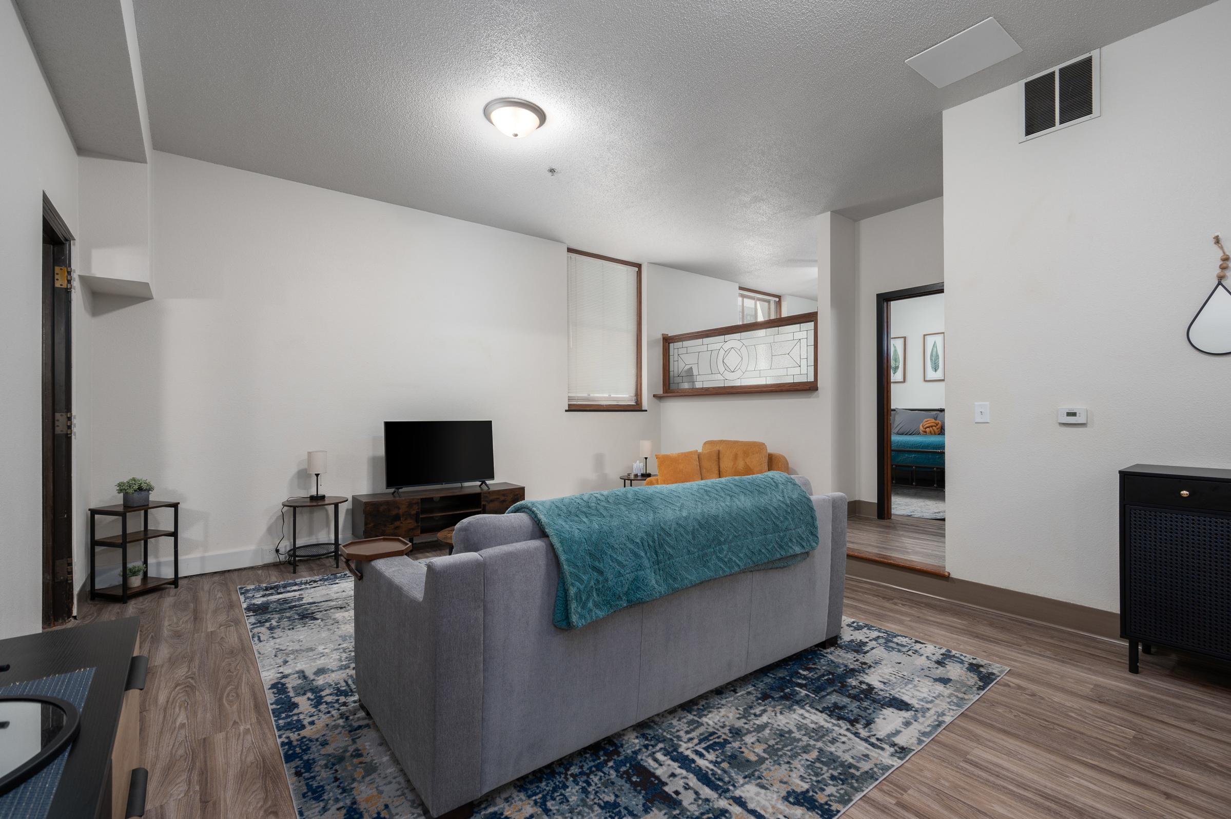 A cozy living room featuring a gray sofa with a blue blanket, an orange armchair, a coffee table, and a TV. The floor is adorned with a large area rug, and the walls are softly colored. A glimpse of another room is visible through an open doorway, adding depth to the space.