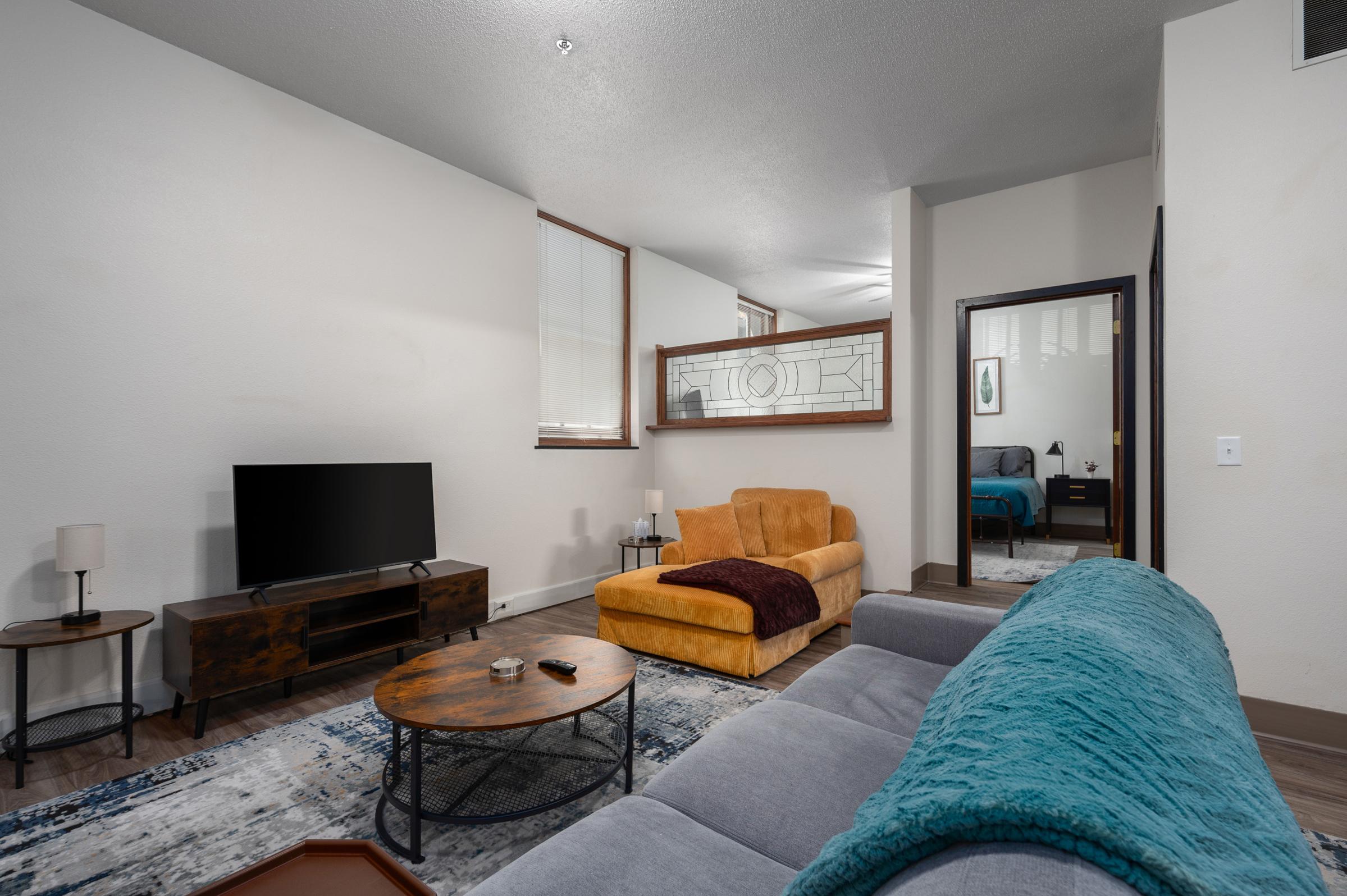 A bright living room featuring a gray sofa with a teal throw blanket, an orange armchair, a round wooden coffee table, and a TV on a low console. A patterned rug covers the floor. In the background, a partition separates the space, and a glimpse of a blue bedroom is visible through a doorway.