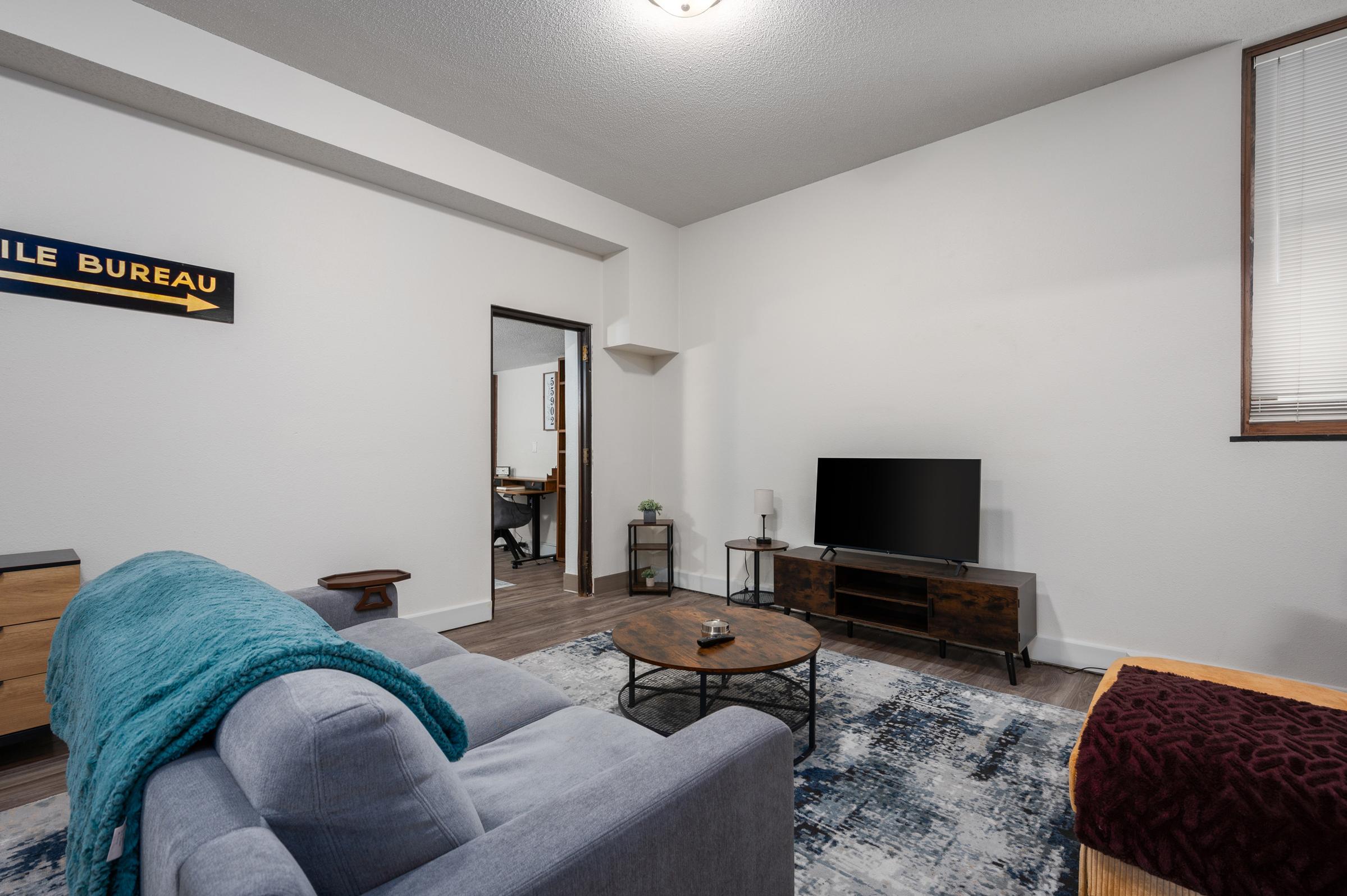 A cozy living room featuring a light gray sofa with a blue throw, a round coffee table, and a dark wooden TV stand with a flat-screen TV. The walls are painted white, and there's a patterned rug on the floor. A small workspace is visible in the background through an open doorway.