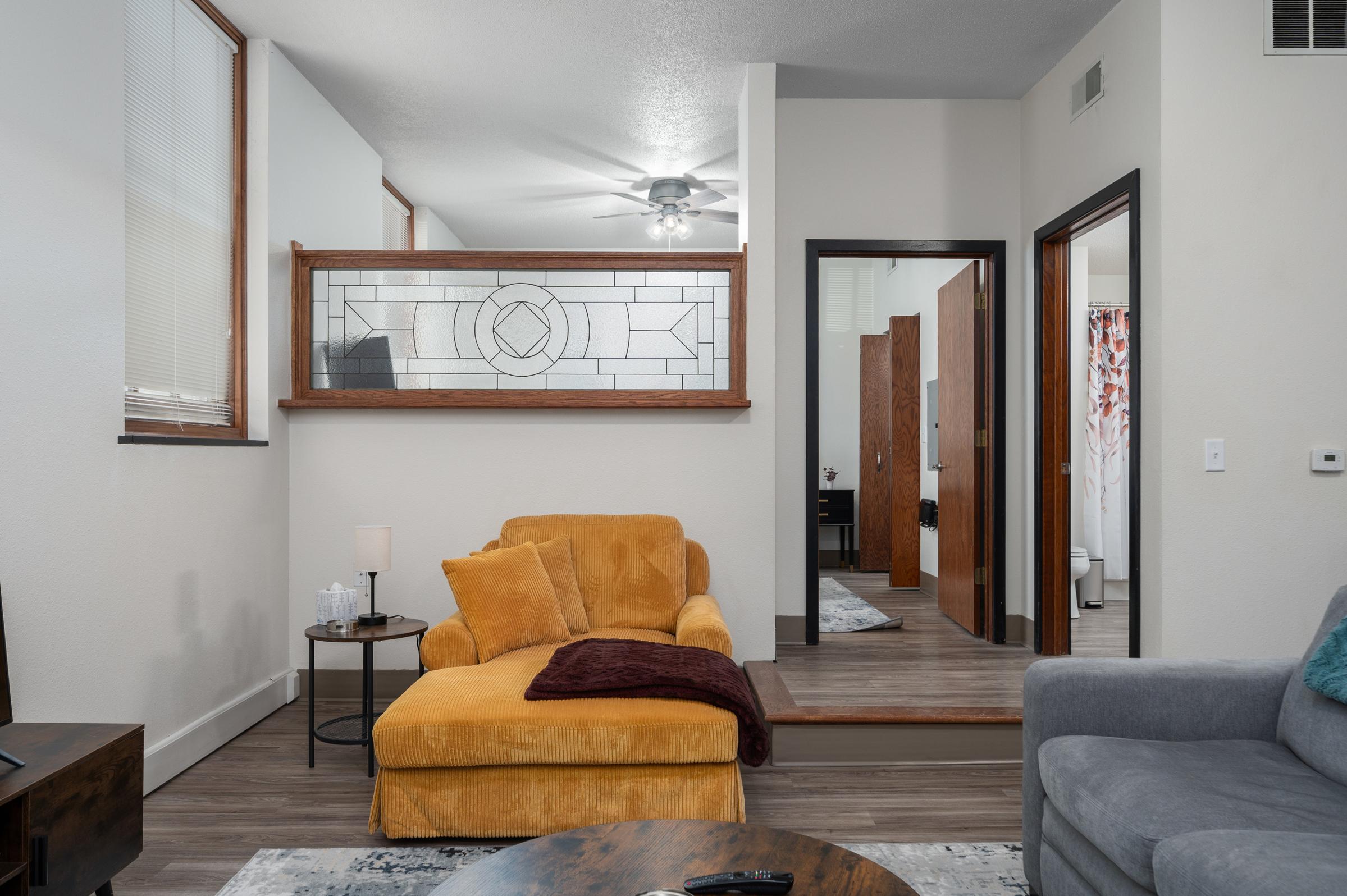 Cozy living space featuring a mustard yellow armchair with a maroon throw blanket, a small lamp on a side table, and a gray sofa. A decorative glass panel separates the seating area from a hallway leading to rooms. Natural light filters through the windows, creating a warm atmosphere.