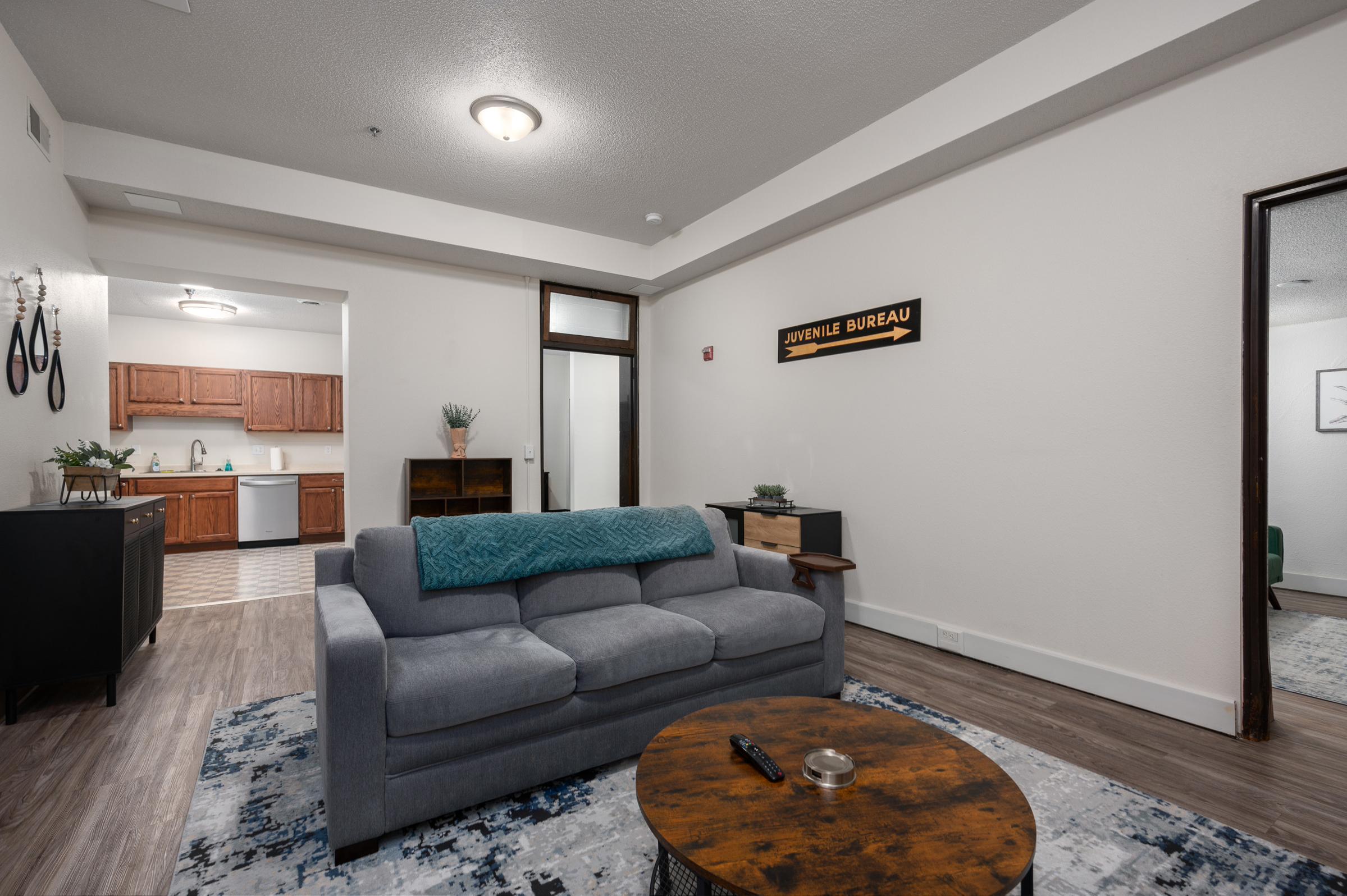 A cozy living room featuring a gray sofa with a blue throw blanket, a round wooden table, and a decorative rug. In the background, there's a kitchenette with wooden cabinets and a sign that reads "JUVENILE BUREAU" on the wall. The space has modern decor and soft lighting.