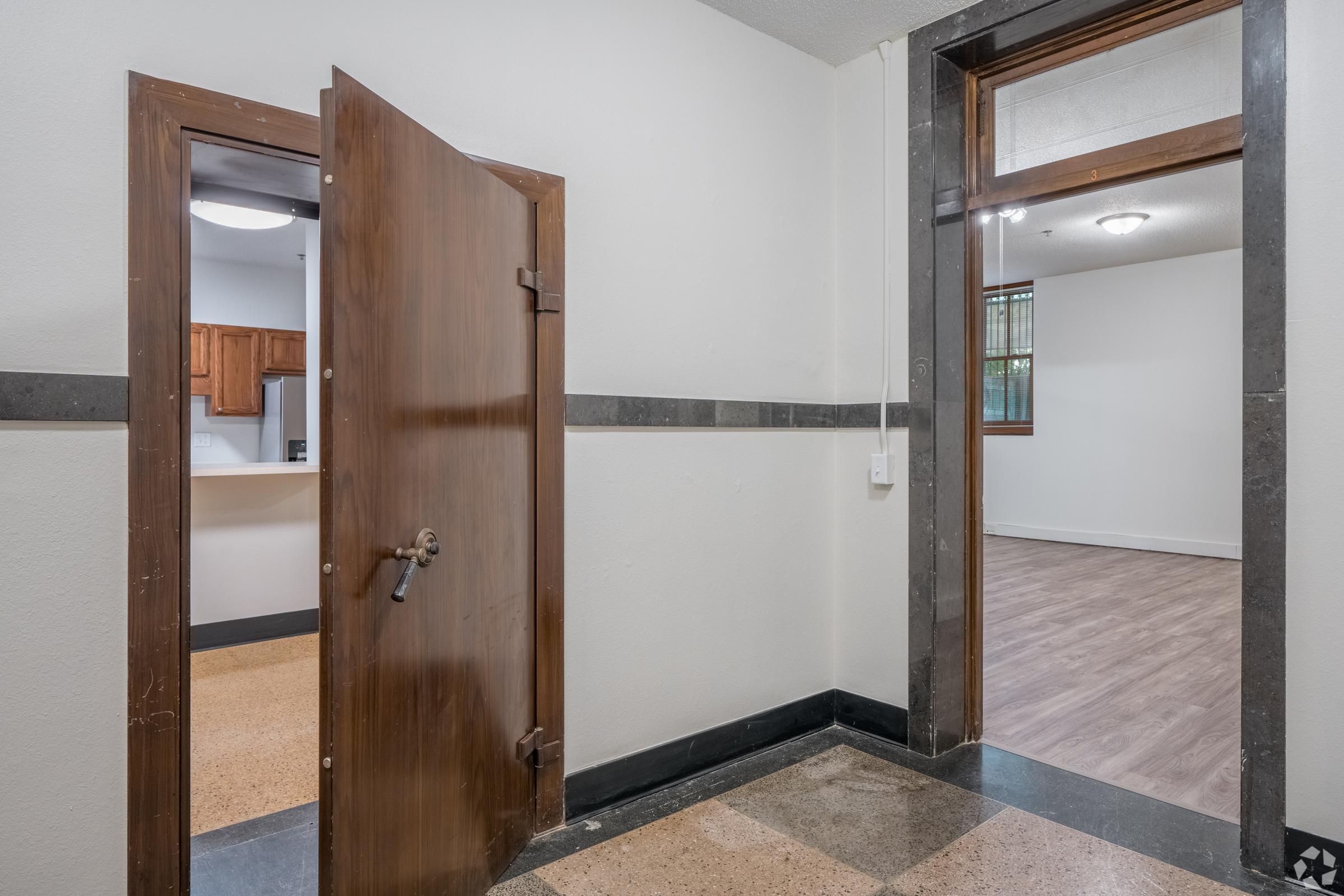 Interior view of a hallway with two open doors. One door leads to a kitchen area with wooden cabinets, while the other opens into an empty room with wood flooring. The walls are neutral-colored, and the flooring features a combination of tiles and polished surfaces.