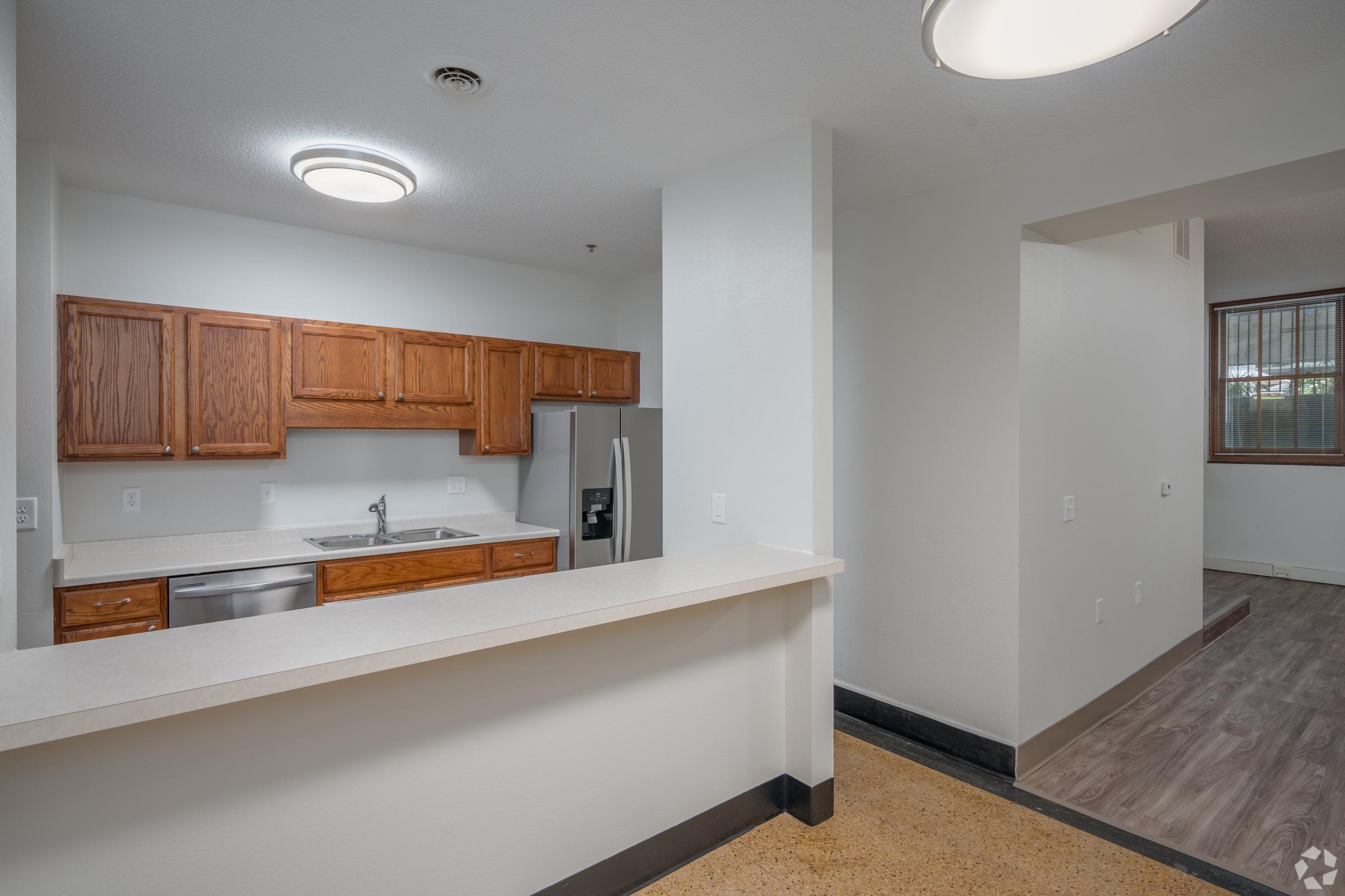 A modern kitchen with wooden cabinets and a countertop, featuring stainless steel appliances including a refrigerator and sink. The open layout connects to a spacious living area with natural light from a large window. The floor has a combination of smooth tiles and laminate.