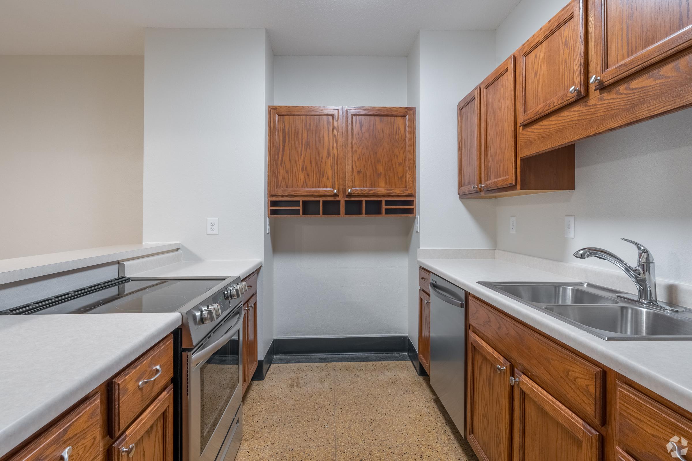 A modern kitchen featuring wooden cabinets, a double sink, stainless steel appliances including a stove and dishwasher, and a light-colored countertop. The space has an open layout with neutral wall colors, providing a clean and inviting atmosphere.