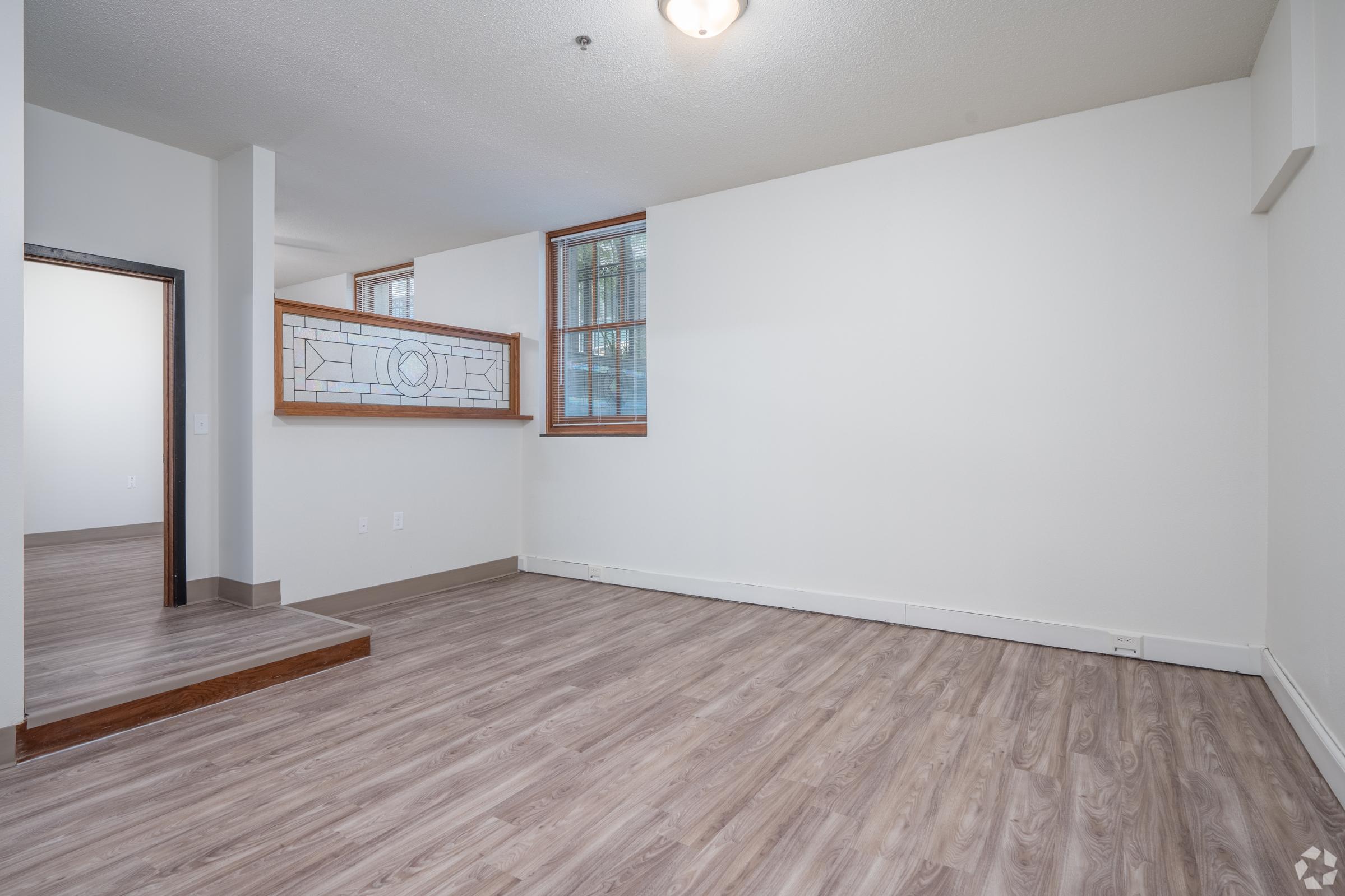 Interior view of a bright, modern living space featuring light-colored walls, large windows, and hardwood flooring. A decorative glass partition separates the main area from another room, adding architectural interest. The space is open and inviting, ideal for various furniture arrangements.