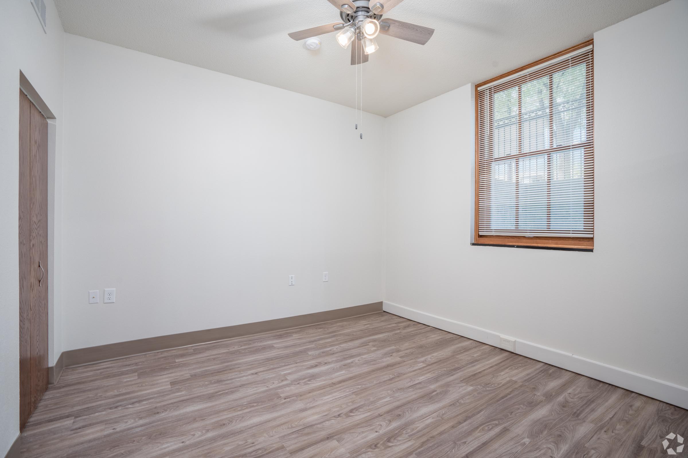 A spacious, empty room featuring light-colored walls, a window with blinds, and a ceiling fan. The floor is made of light brown wood laminate, and there are two electrical outlets visible on the wall. The overall feel is bright and open, making it suitable for various uses.