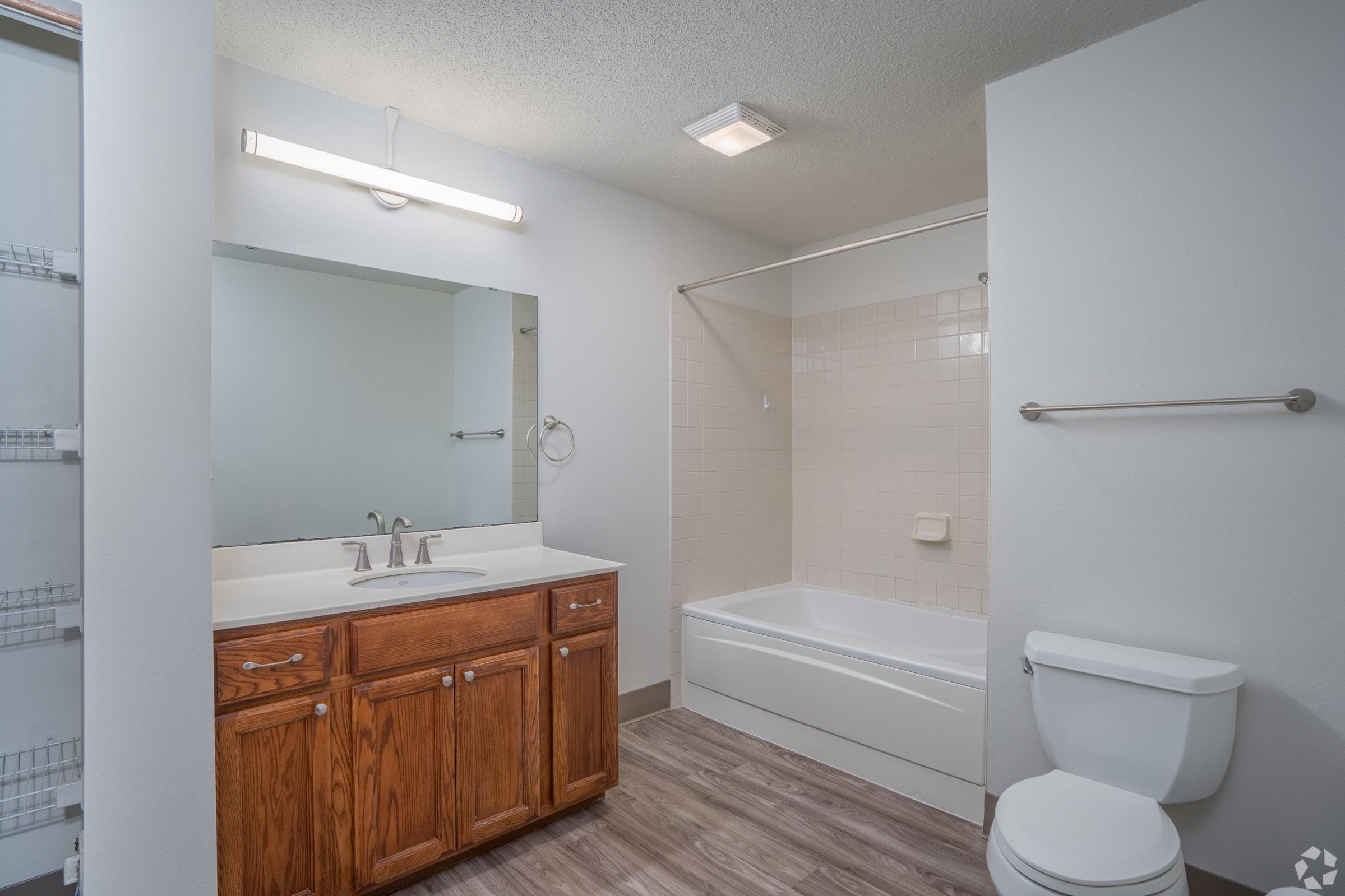 A bathroom featuring a wooden vanity with dual sinks, a large mirror above, a bathtub with a shower curtain, a toilet, and laminate flooring. Natural light from a ceiling fixture illuminates the space, which has neutral-colored walls and is organized and clean.