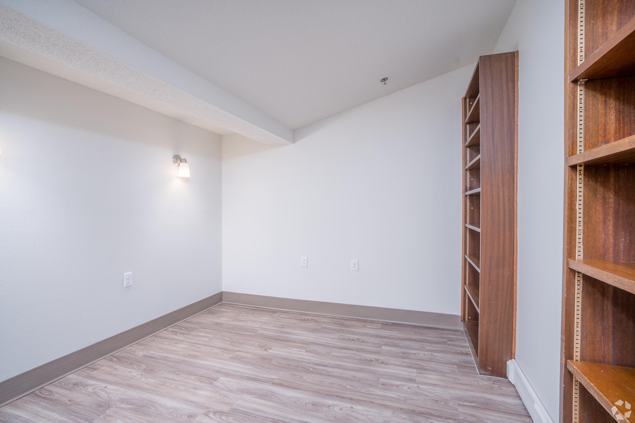 A small, empty room with light-colored walls and a wooden floor. There is a wall-mounted light fixture on the left and a built-in bookshelf on the right. The space is designed for potential use as an office or storage area.