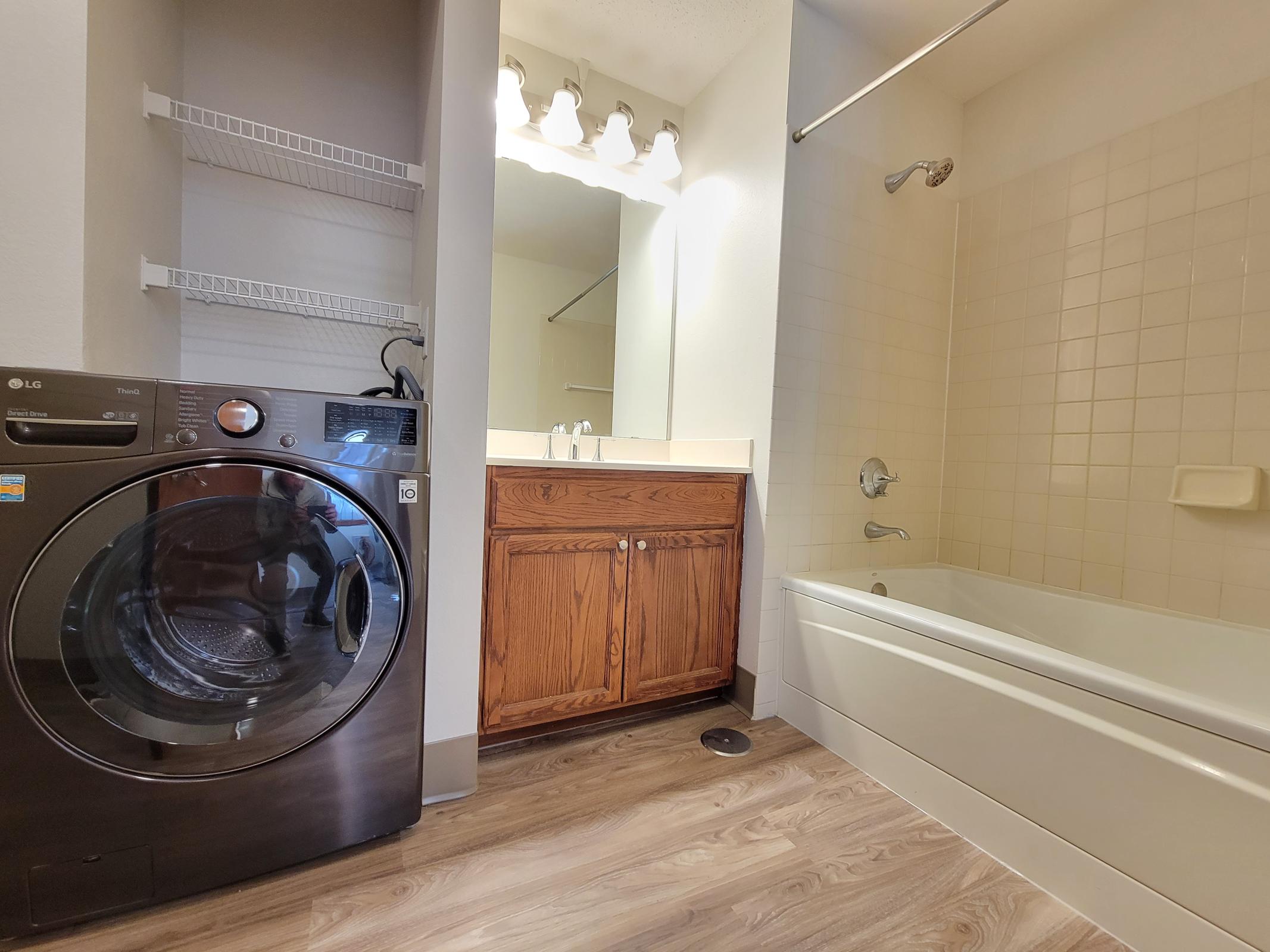 A modern laundry area featuring a black front-loading washing machine next to a bathroom. The bathroom includes a bathtub, a sink with a wooden cabinet, and bright lighting from above. White tiles and light wood flooring enhance the contemporary design.