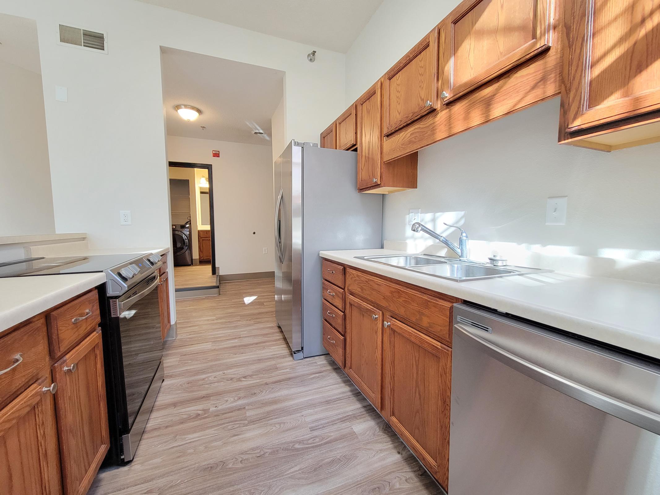 A modern kitchen featuring wooden cabinets, stainless steel appliances, and a double sink. The space includes a refrigerator, dishwasher, and stove, with light-colored countertops. Sunlight streams in through a window, highlighting the wood-like floor. The kitchen opens to a hallway leading to additional areas.