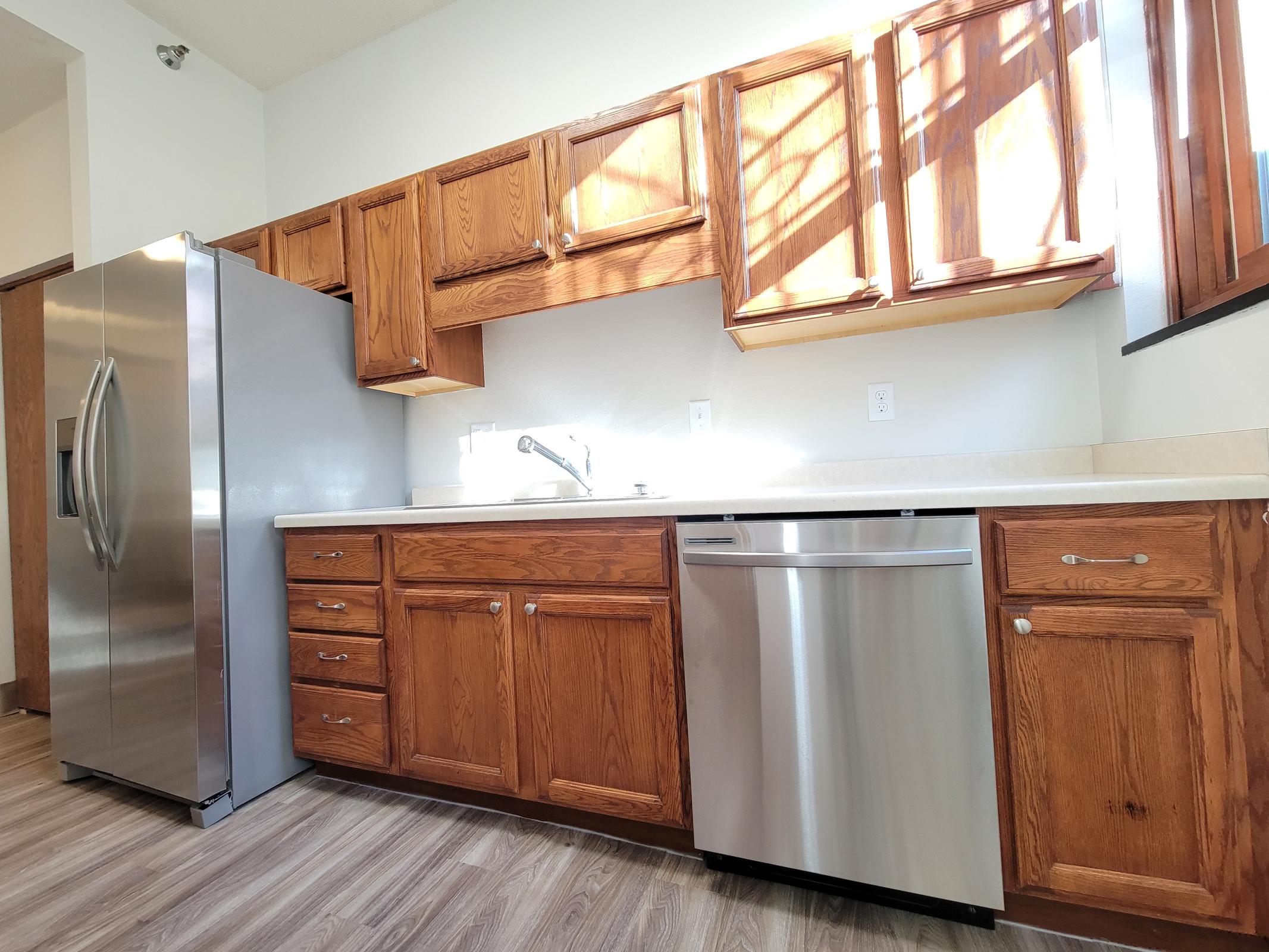 A modern kitchen featuring wooden cabinets, a stainless steel refrigerator, a dishwasher, and a double sink. Natural light streams in from a window, illuminating the countertops and cabinetry. The floor is designed with laminate wood planks for a warm, inviting look.