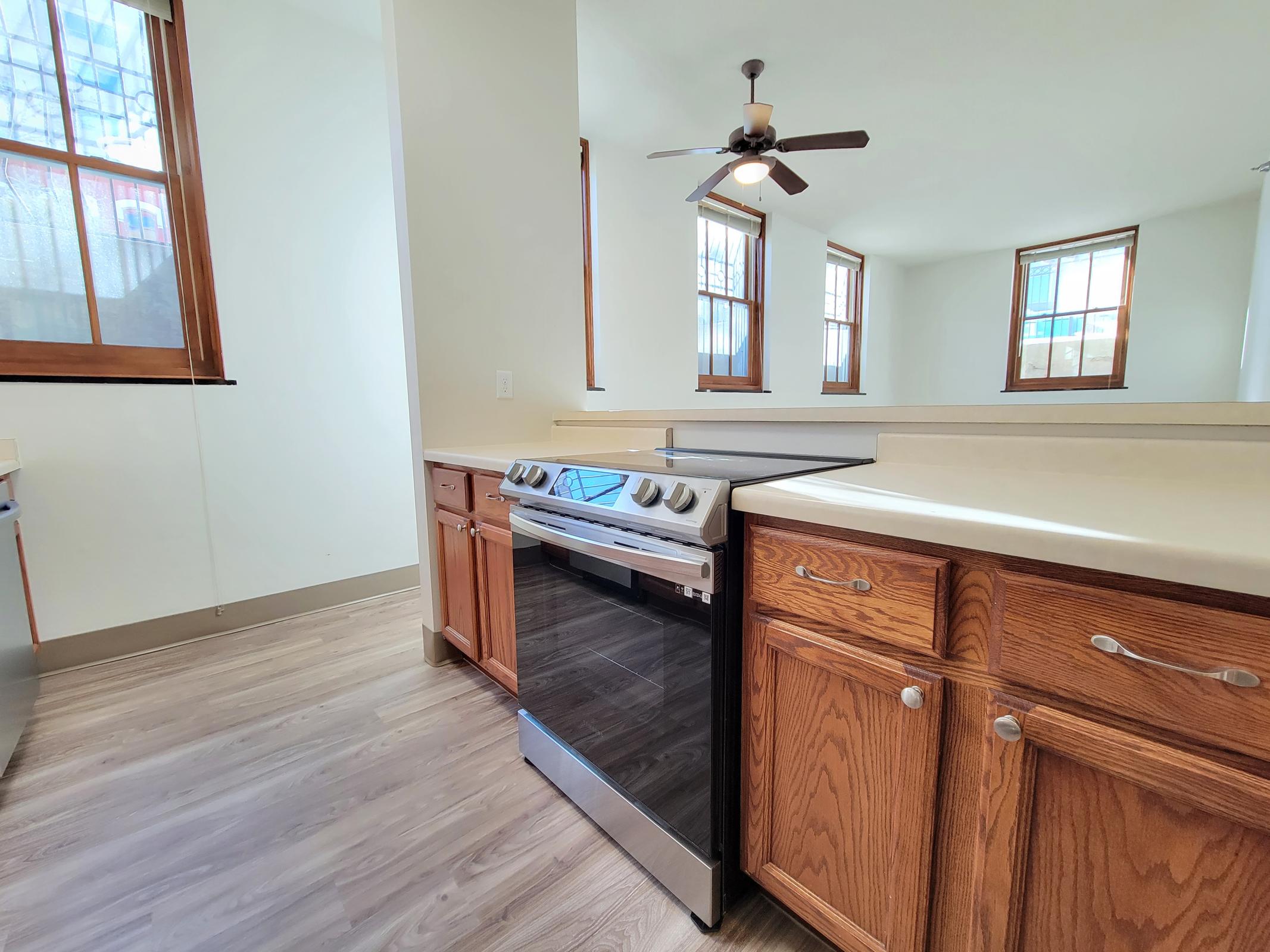 A modern kitchen featuring an oven with a sleek glass surface, wooden cabinets, and a light-colored countertop. Natural light streams in through large windows, illuminating the space. The flooring is a light laminate, adding to the contemporary feel. A ceiling fan is visible, enhancing airflow in the room.