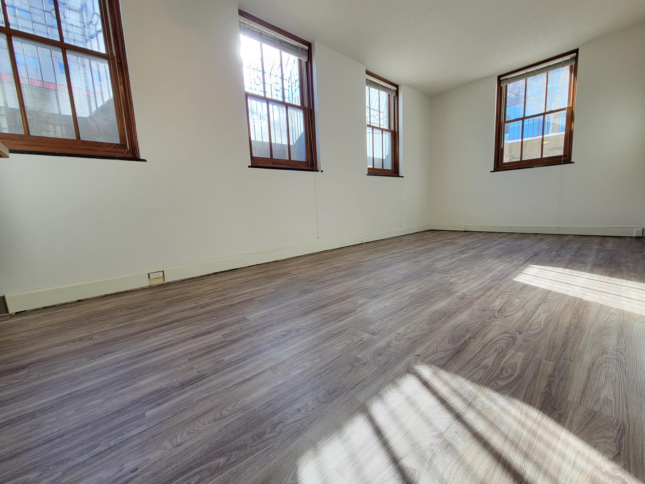 A spacious, empty room with light-colored hardwood flooring. Two large windows with wooden frames allow natural light to enter, casting shadows on the floor. The walls are painted white, creating a bright and airy atmosphere. The space is uncluttered and ready for furniture.