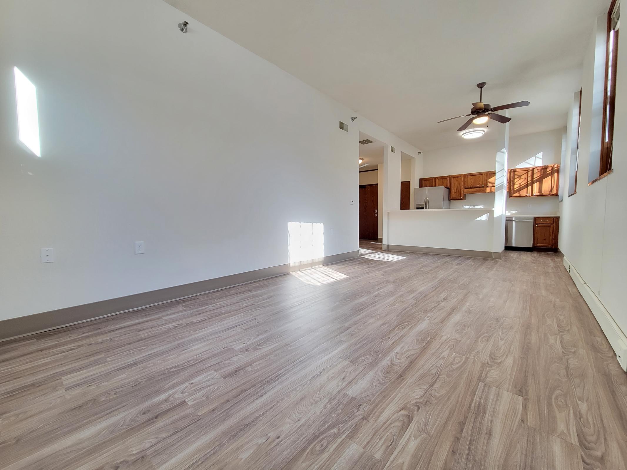 Spacious living area featuring light-colored laminate flooring, a ceiling fan, and sunny windows. The open layout includes a kitchen area with wooden cabinets, a countertop, and a nearby dining space. Natural light streams in, creating a warm and inviting atmosphere.