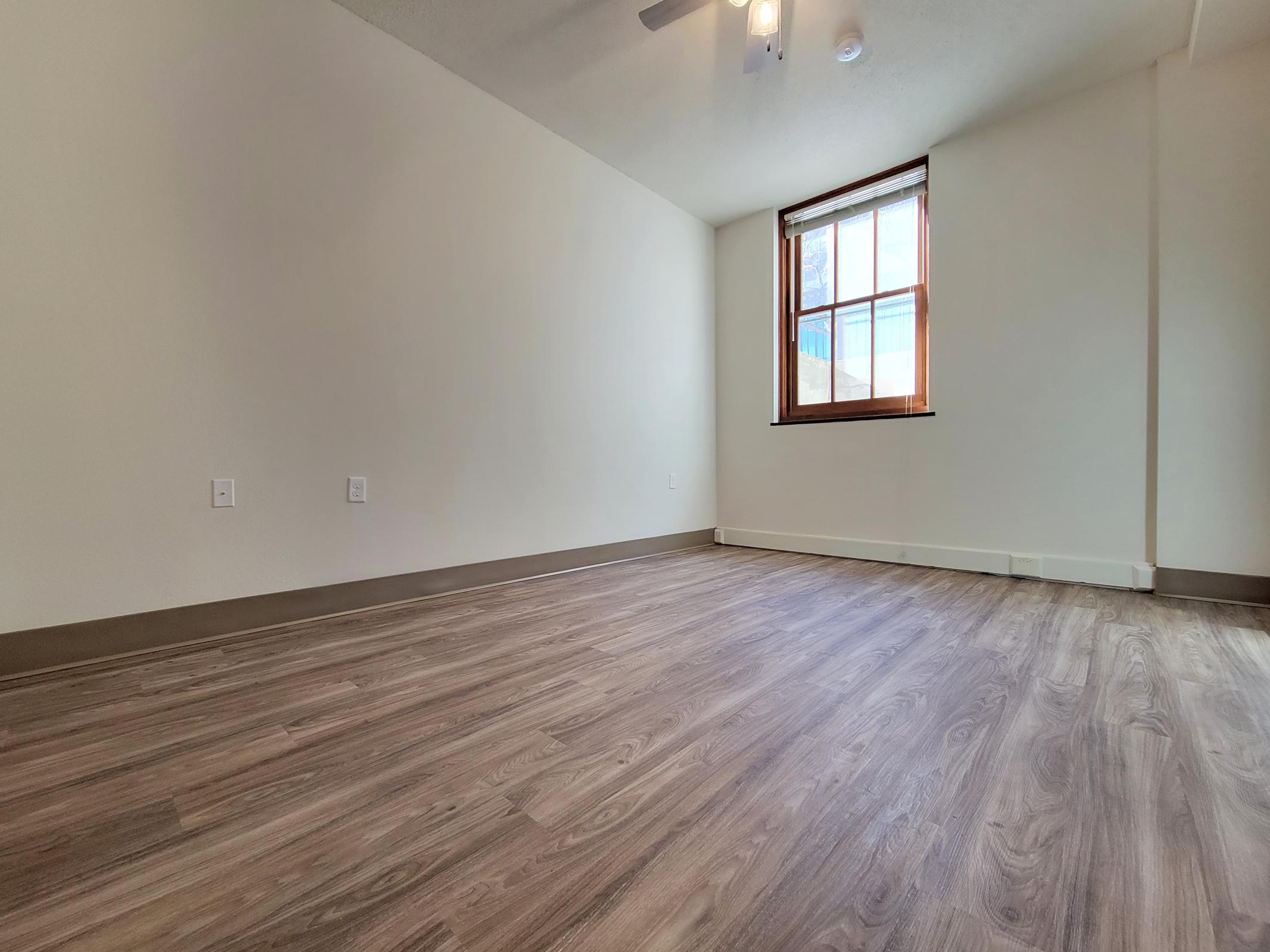 A spacious, empty room featuring light-colored walls and a wooden floor. There is a window on one wall allowing natural light to enter, and a ceiling fan is visible. The overall atmosphere is bright and minimalist, ideal for various uses.