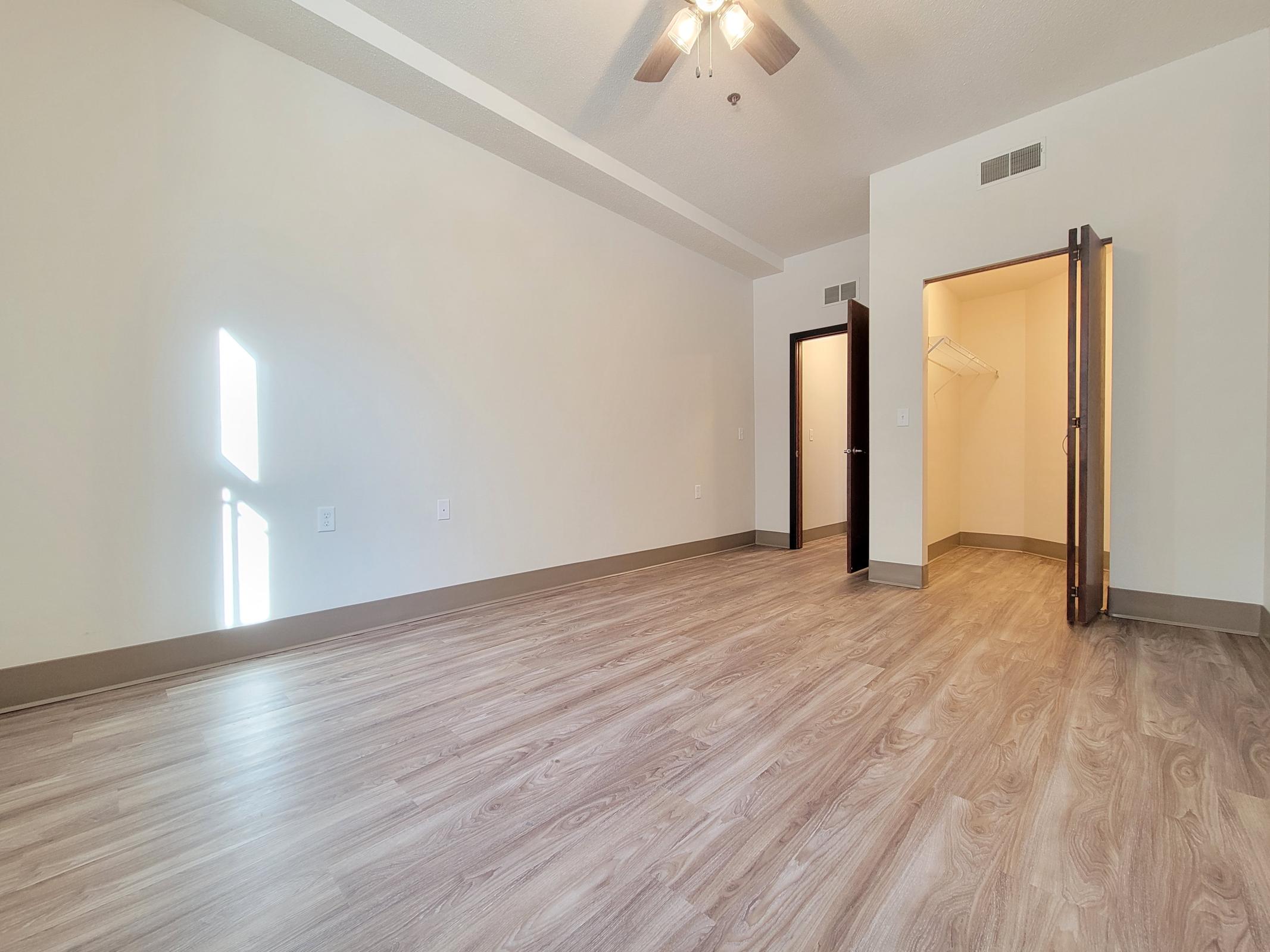 A spacious, empty room with light-colored walls and laminate flooring. A ceiling fan is visible, and there is a closet on the right side featuring a door that is slightly open. Natural light is streaming in from a window, creating a bright atmosphere.