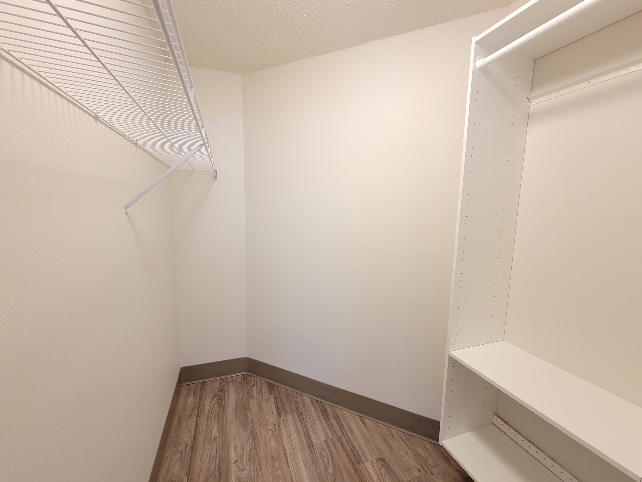 A spacious and empty closet featuring light-colored walls, a wooden floor, and a wire shelf on one side. The opposite side has a white built-in shelf unit, creating a clean, organized space ready for storage.