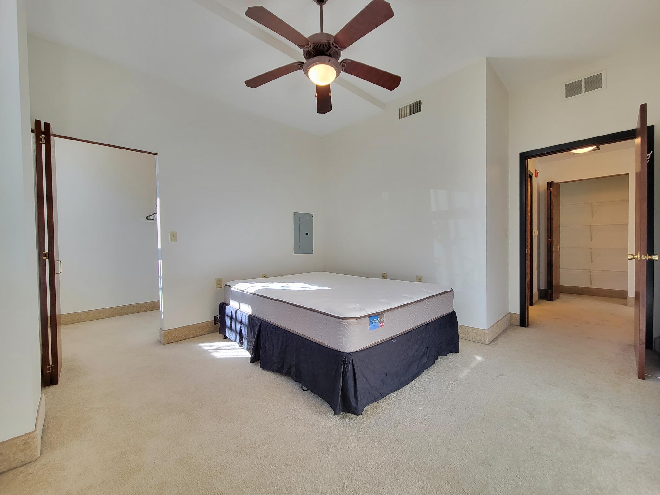 Spacious bedroom with light-colored walls and carpet. A ceiling fan hangs above an unmade bed with a dark bed skirt. There are two doorways, one leading to a closet and another to a small hallway, allowing natural light to enter the room.
