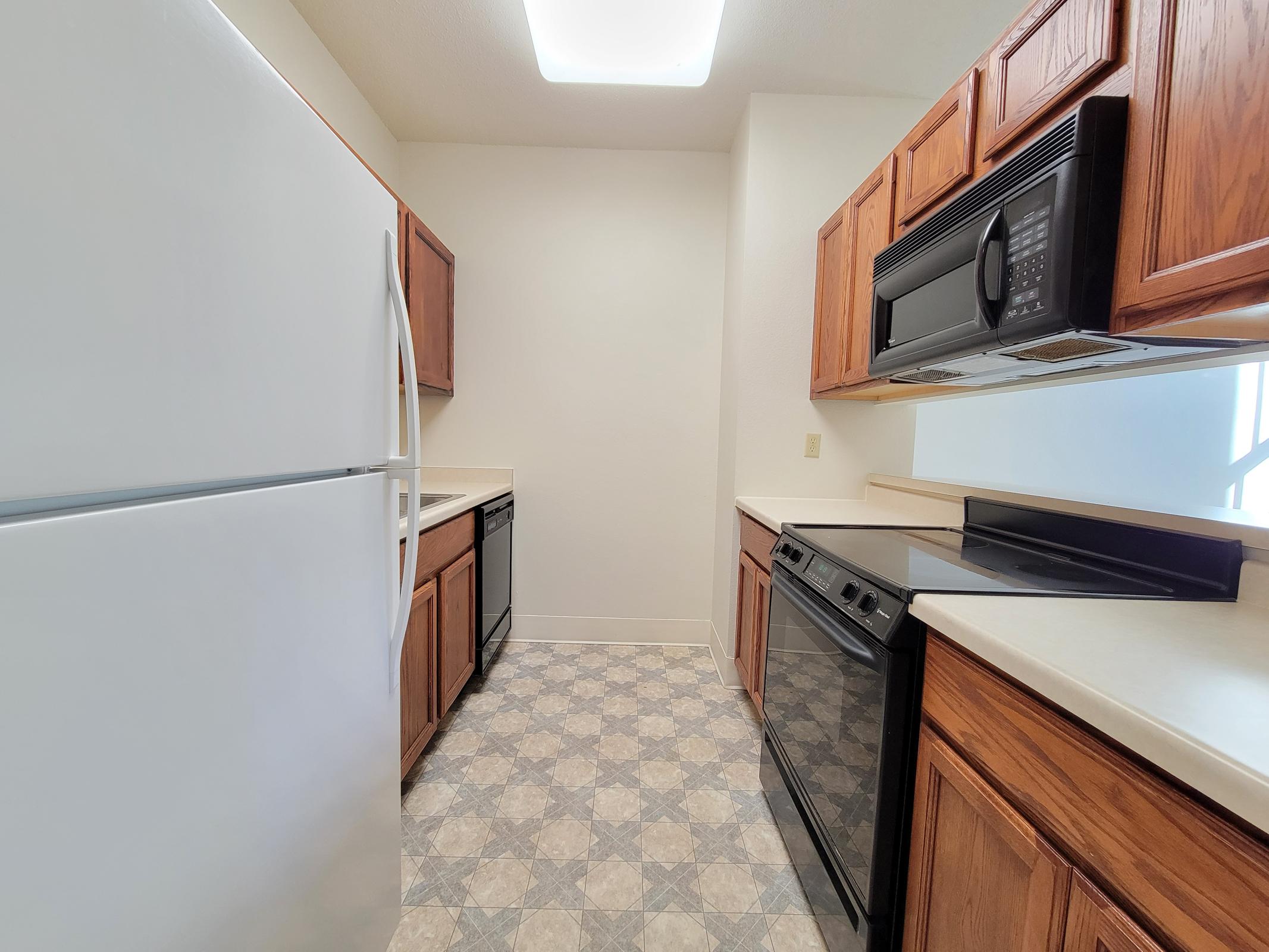 A modern kitchen featuring wooden cabinetry, white countertops, and gray tiled flooring. The kitchen includes a white refrigerator, a black stove, and a black dishwasher. Ample counter space provides a functional layout with good lighting from a window. Overall, a clean and organized look.