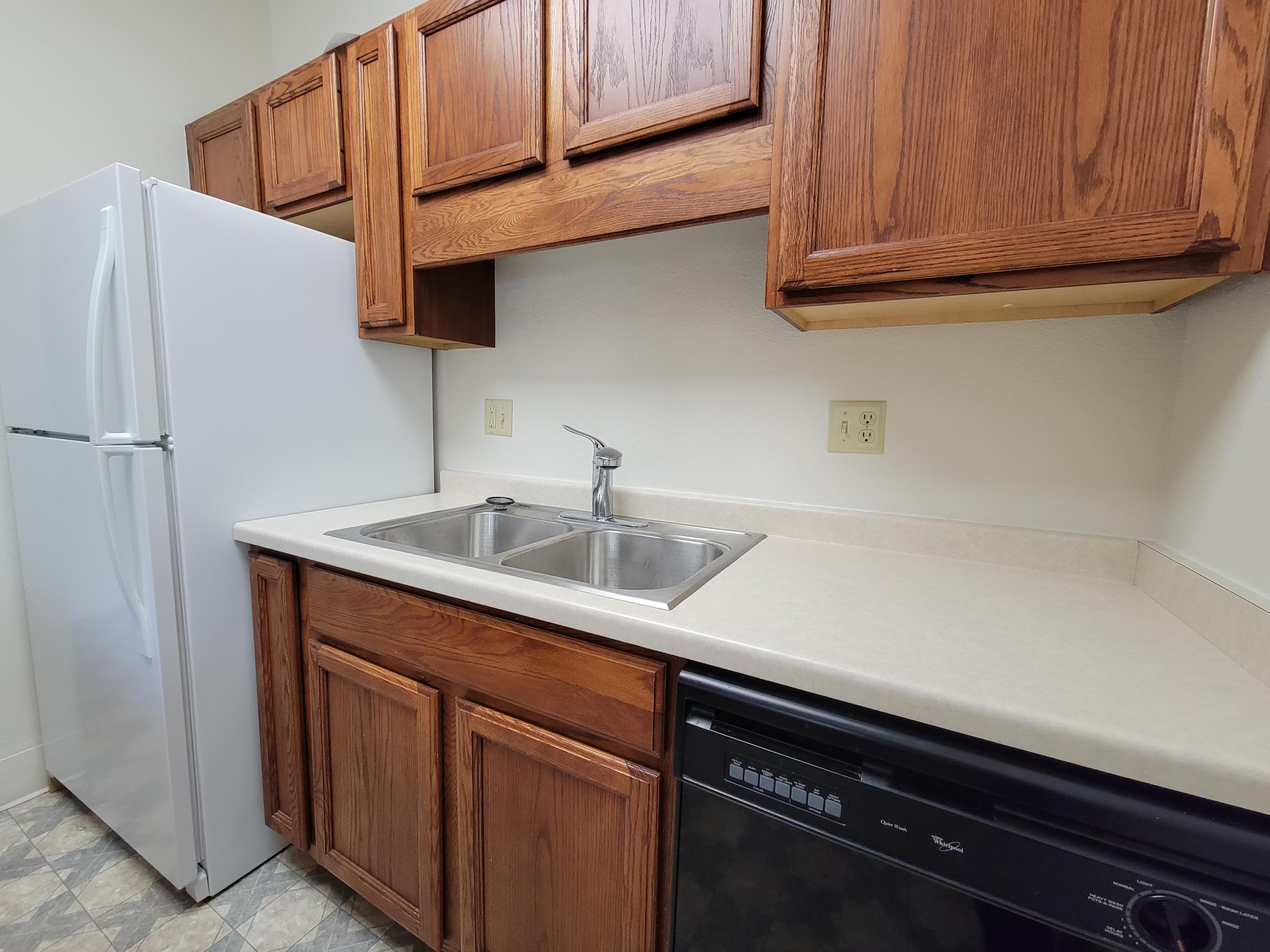 A small kitchen featuring wooden cabinets, a double sink, a white refrigerator, and a black dishwasher. The countertop is light-colored, and the floor has a patterned design. The walls are plain and light-colored, creating a simple, functional space.