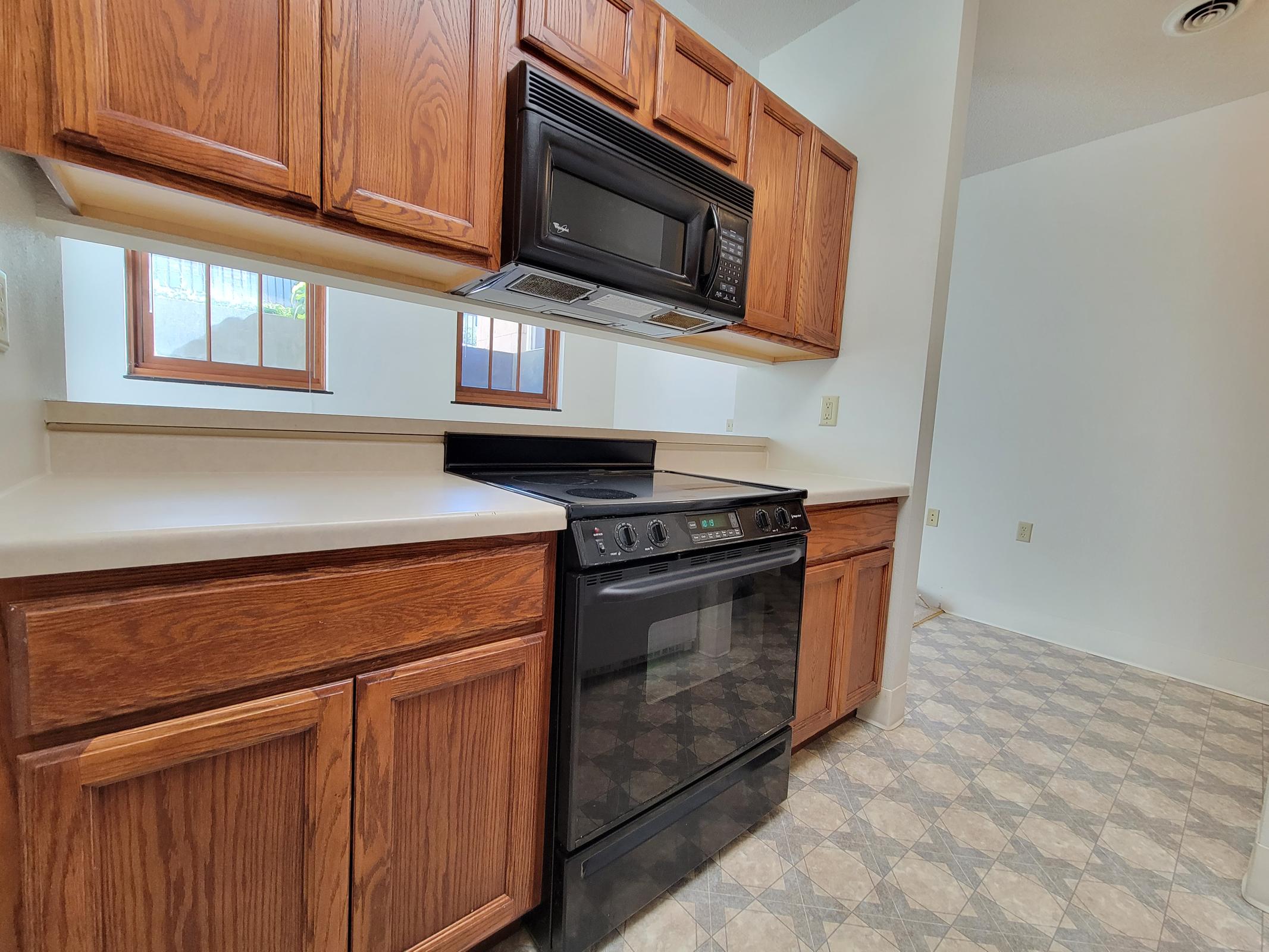 A modern kitchen featuring wooden cabinets, a black microwave, and an oven. The countertop is light-colored, and the floor has a patterned vinyl design. Natural light comes through the windows, providing a bright and inviting space. The kitchen is clean and organized, with ample storage.