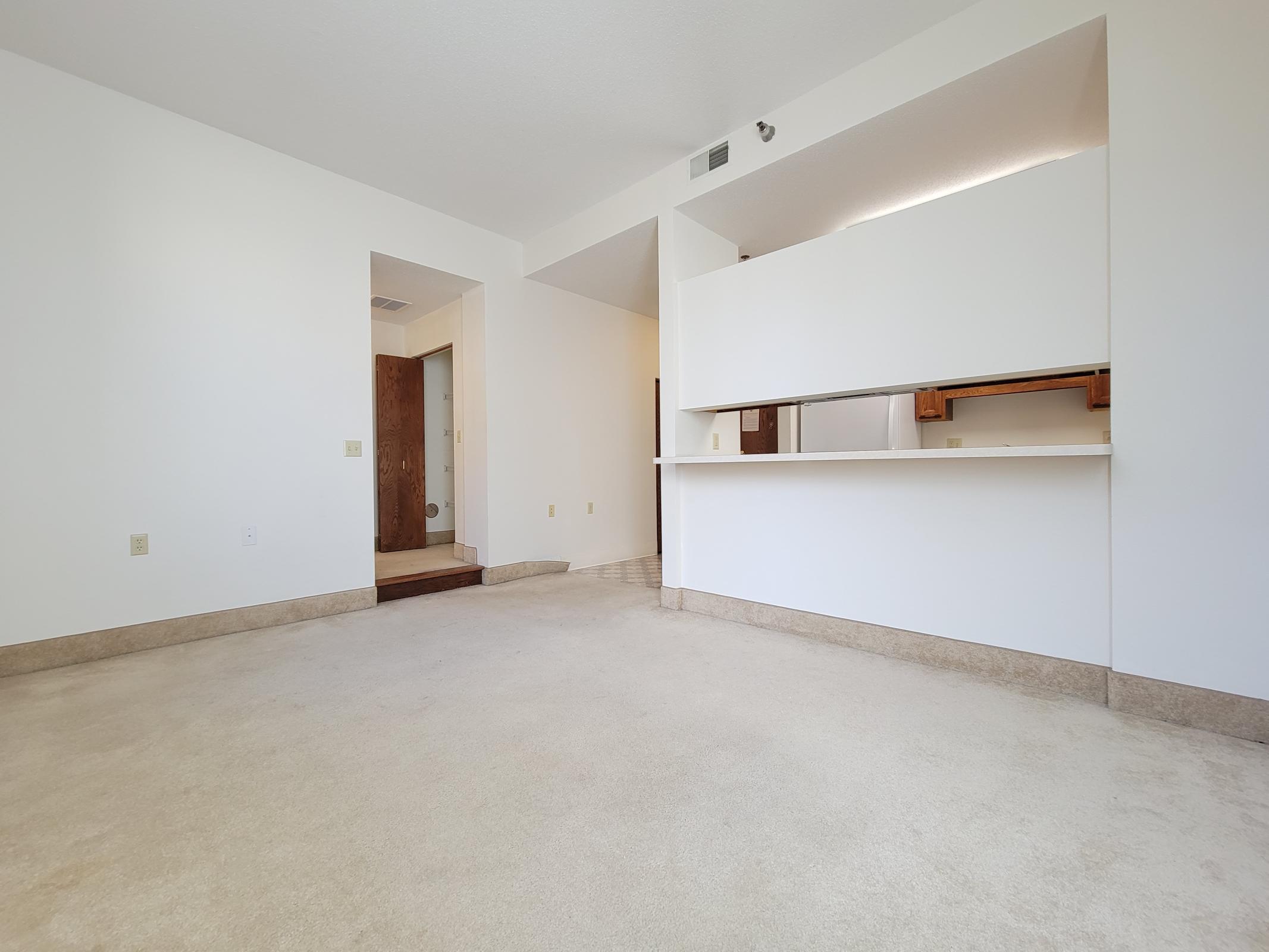 A spacious, empty room featuring beige carpet flooring, white walls, and an open area leading to a kitchen. A doorway is visible on the left, possibly leading to a bathroom or hallway. The room has a bright, neutral ambiance with natural light coming in.