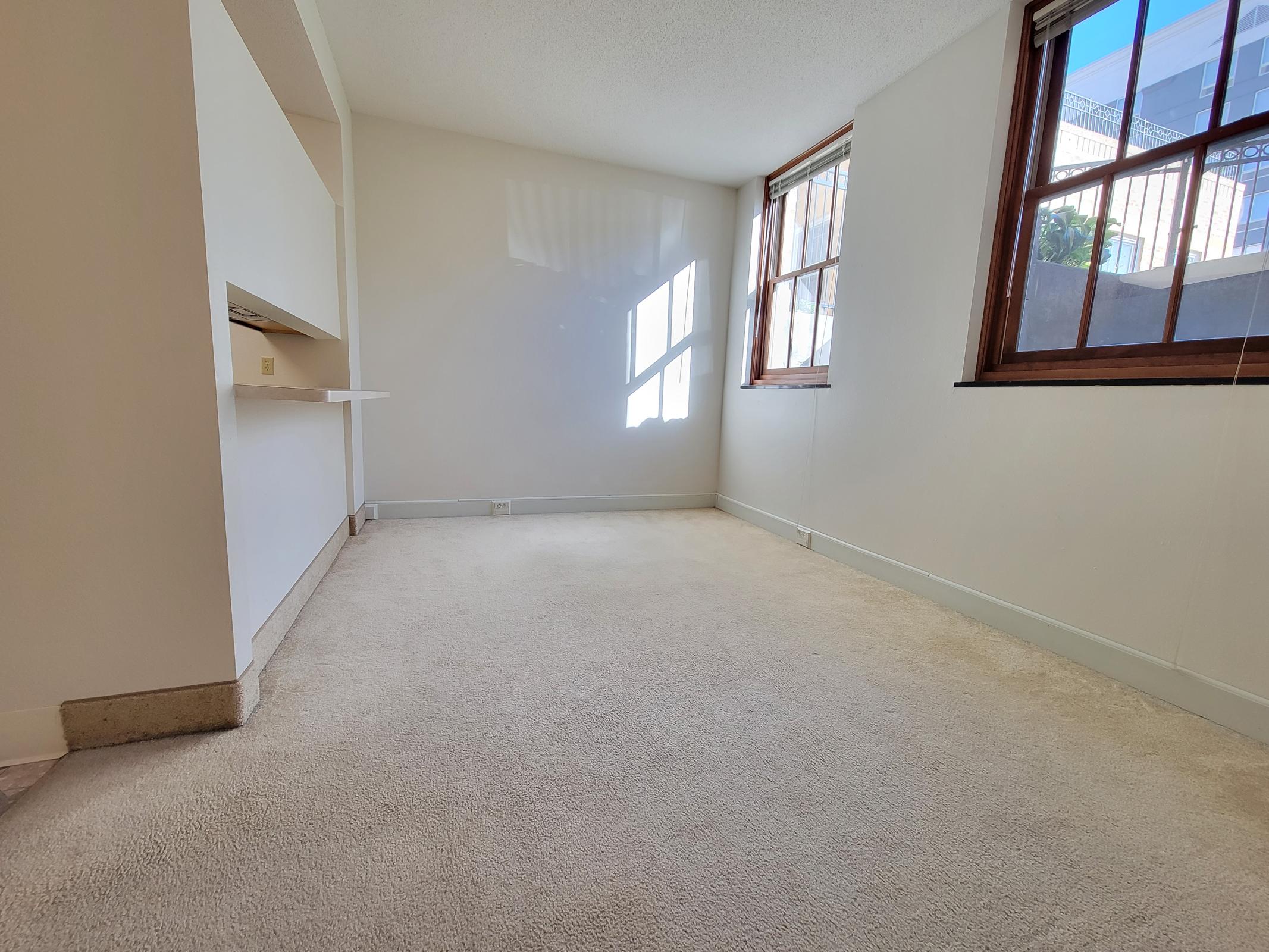 A spacious, empty room with light beige carpet, white walls, and two large windows allowing natural light. A small ledge is visible near the far wall. The room has a minimalistic appearance, suggesting it is ready for furniture or decoration.