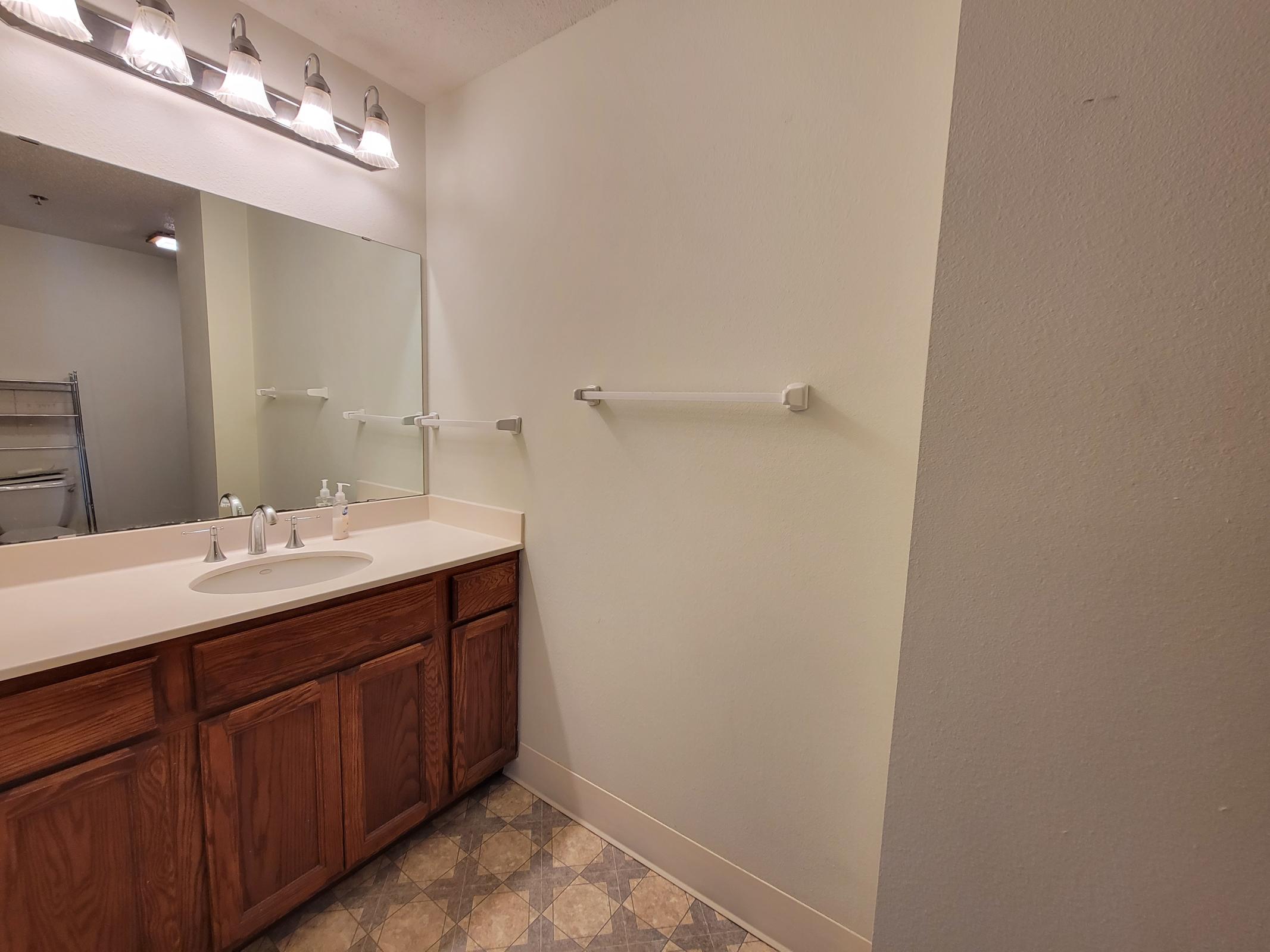 A clean, modern bathroom featuring a light-colored vanity with a double sink and wooden cabinetry. A large wall mirror is above the sink. The walls are a neutral shade, and there's a towel bar on the side wall. The floor has a patterned tile design, creating a spacious and bright atmosphere.