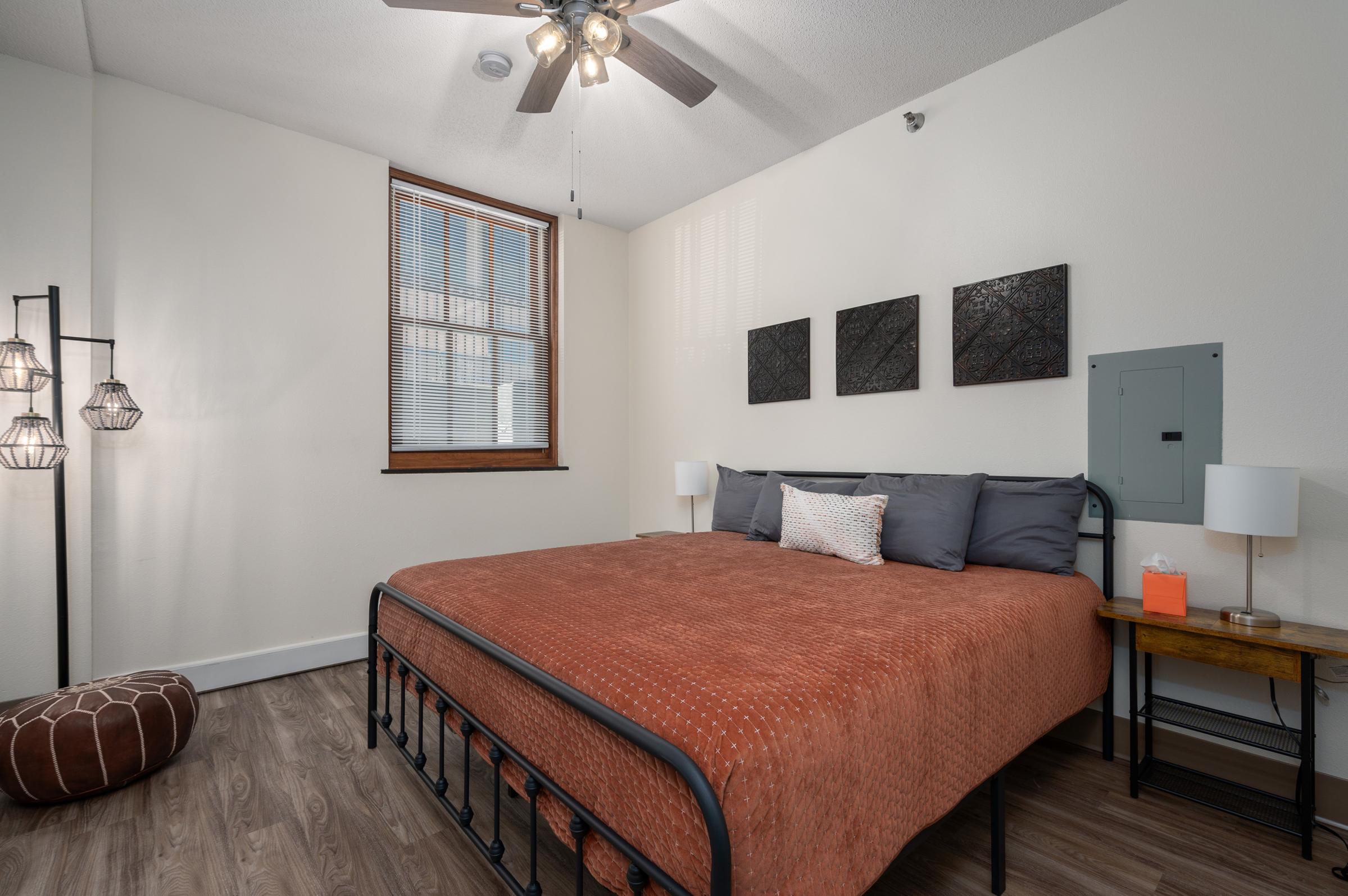 Cozy bedroom featuring a large orange bedspread on a black metal frame, two bedside lamps, a wooden nightstand, and a decorative pouf. The room has a light-colored wall with three framed artworks and a ceiling fan. A window brings in natural light.